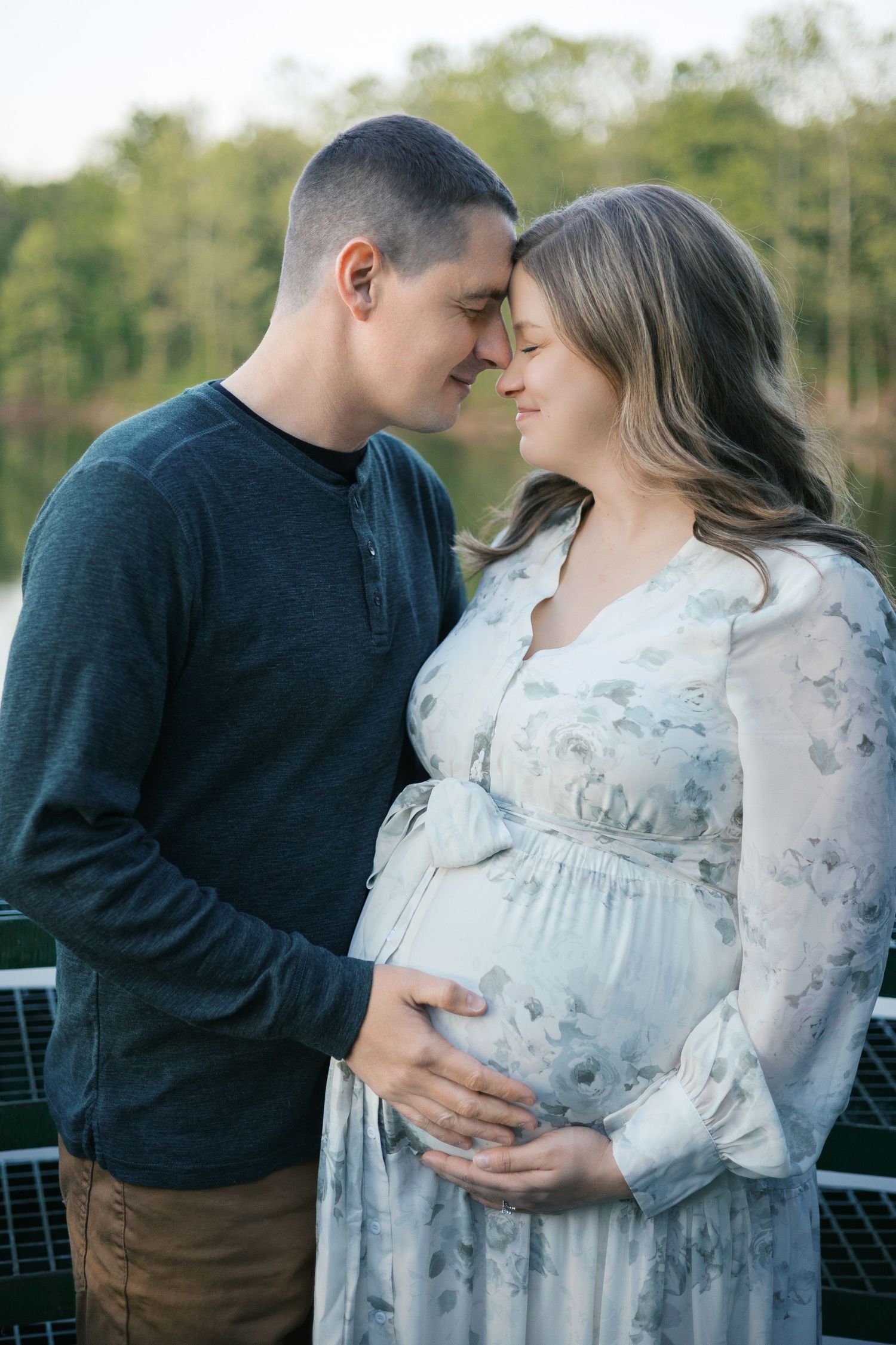 Couple sharing a tender moment during maternity photo shoot outdoors, woman in floral dress cradling baby bump.
