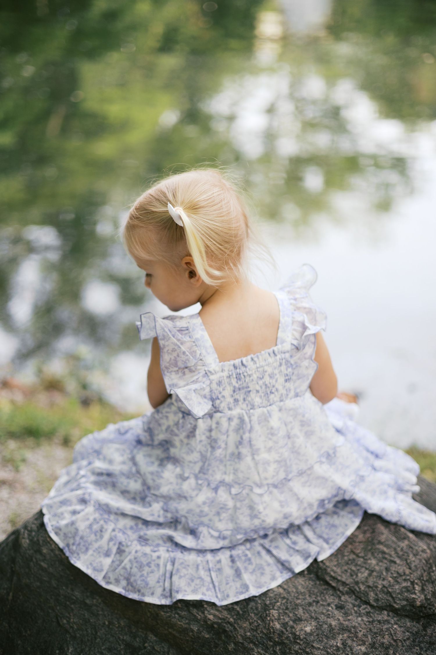 Young girl in blue floral dress sits on rock by pond during Holcomb Gardens family session by Katy Rose Photography.