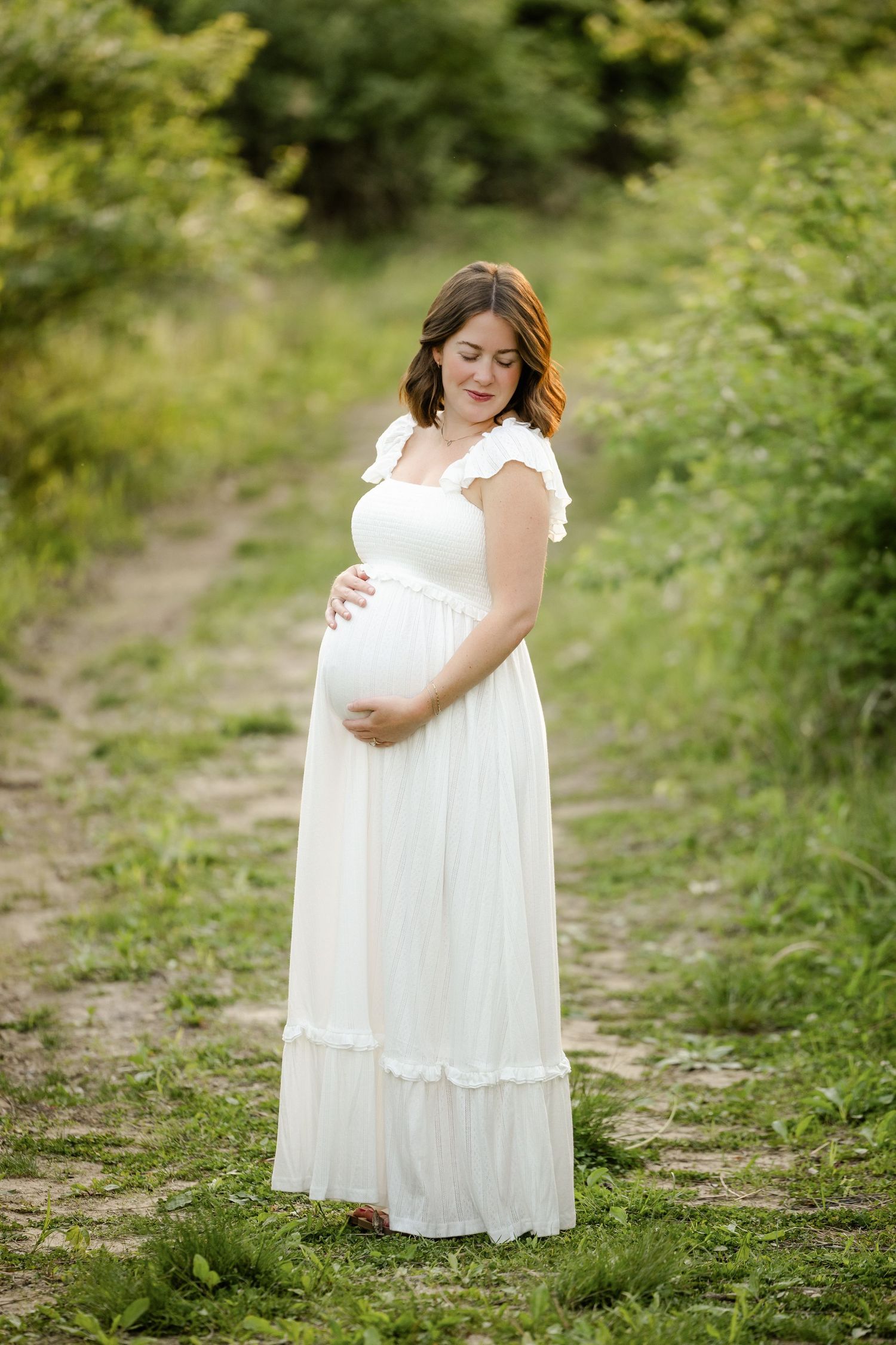 Pregnant woman in white dress cradling baby bump on a scenic garden path during Indianapolis Maternity Portraits by Katy Rose Photography.