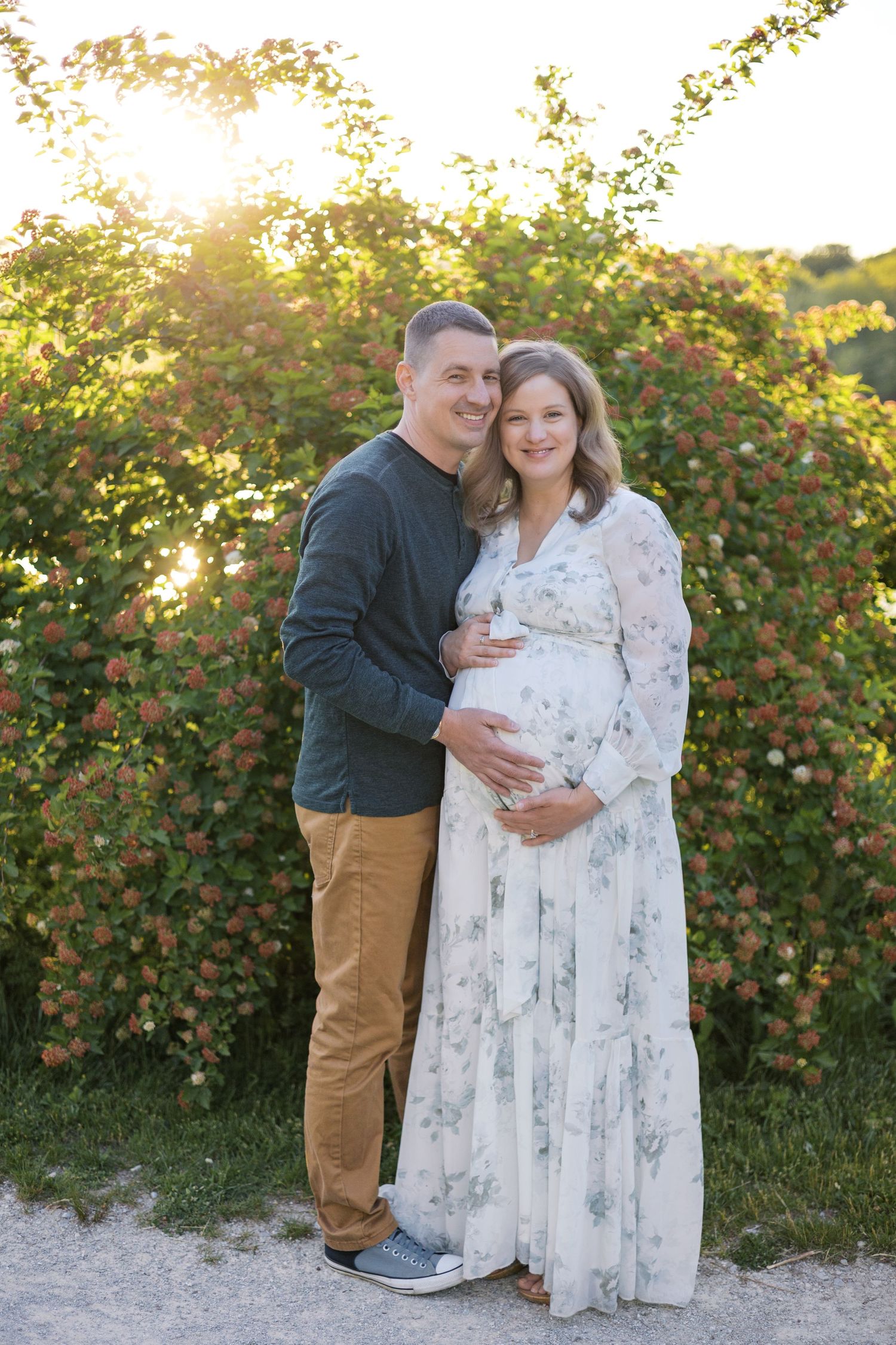 Smiling couple poses before flowering bushes at golden hour, pregnant woman in white floral dress, maternity photo.
