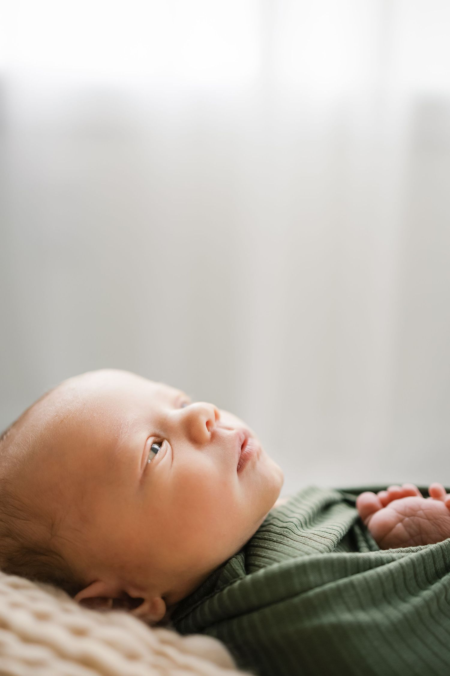 Newborn baby wrapped in a green blanket gazing upward with soft natural light in the background.