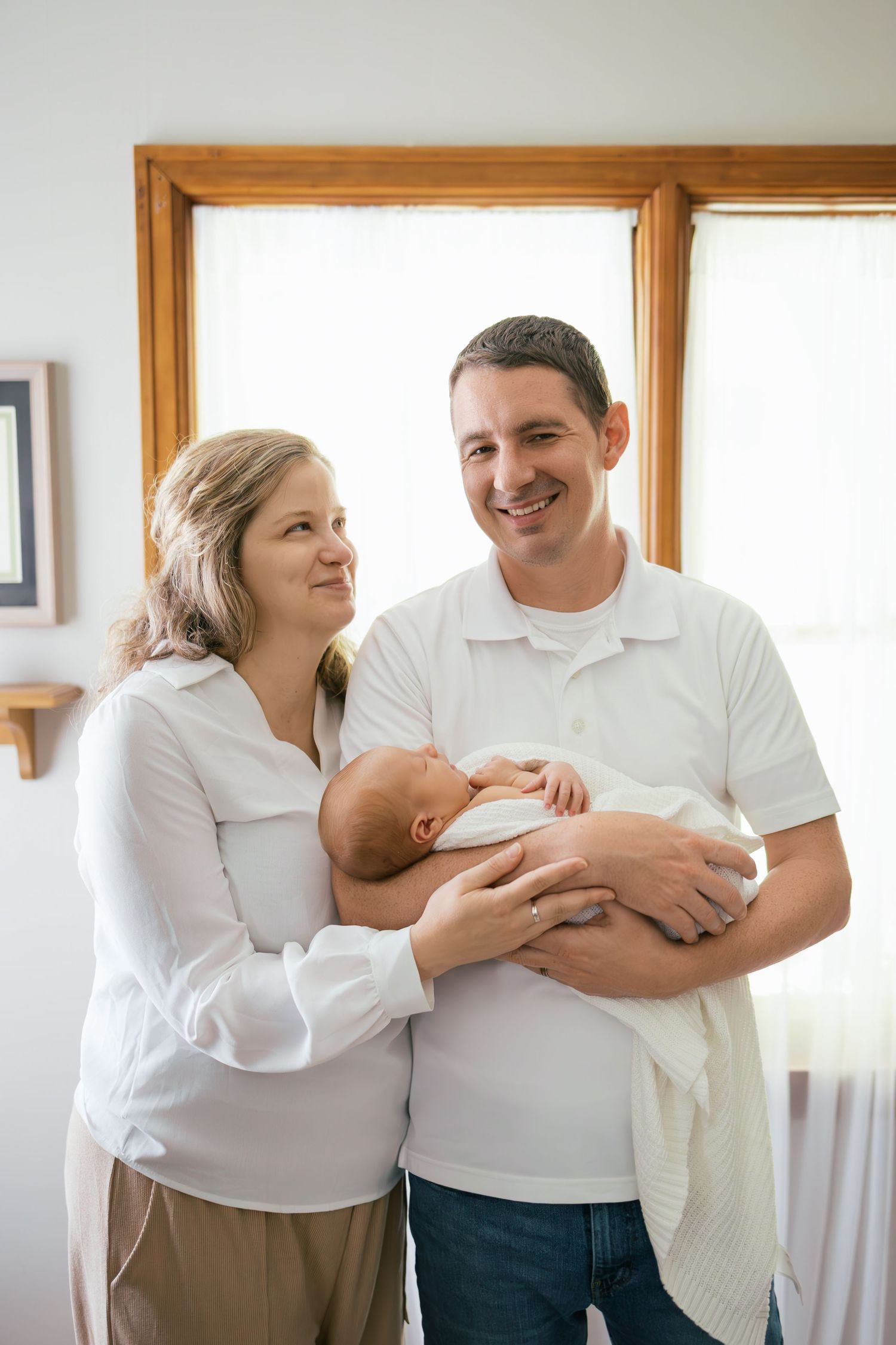 Happy couple in white shirts smiling while holding their newborn baby in a bright home nursery.