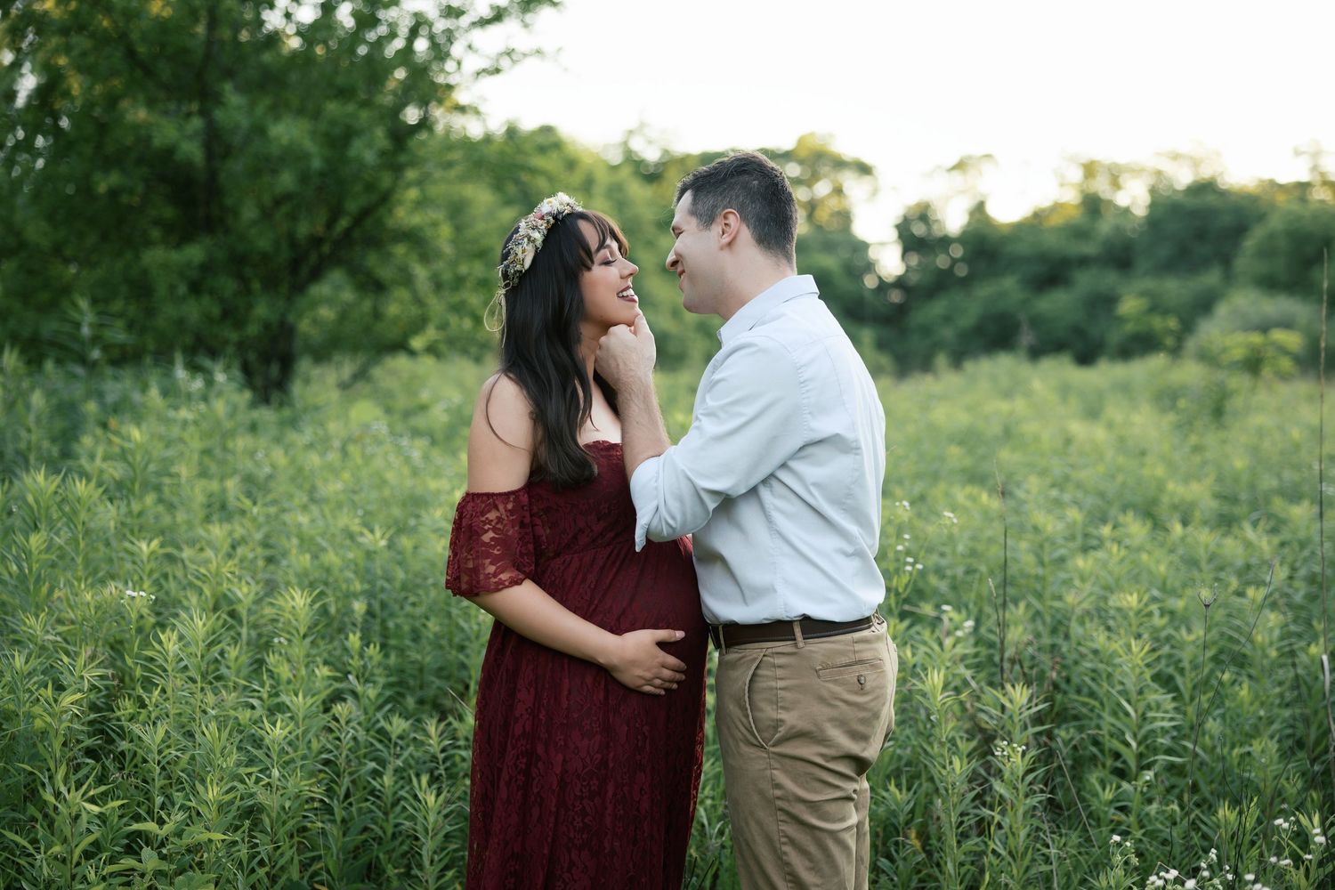 Couple sharing a tender moment in a lush green field, woman in red maternity gown with floral crown.