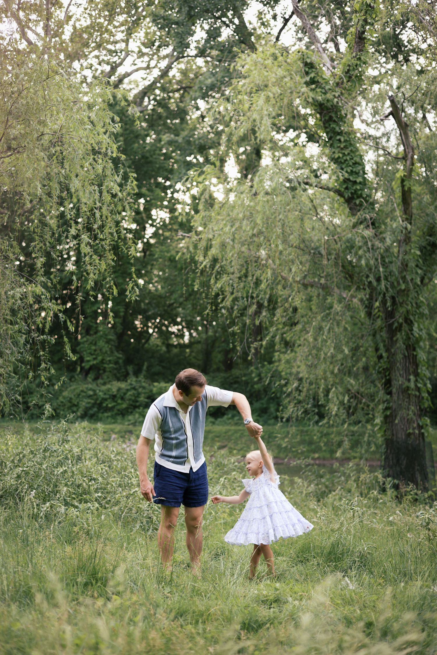 Father and toddler daughter dancing together in a green meadow surrounded by tall trees.