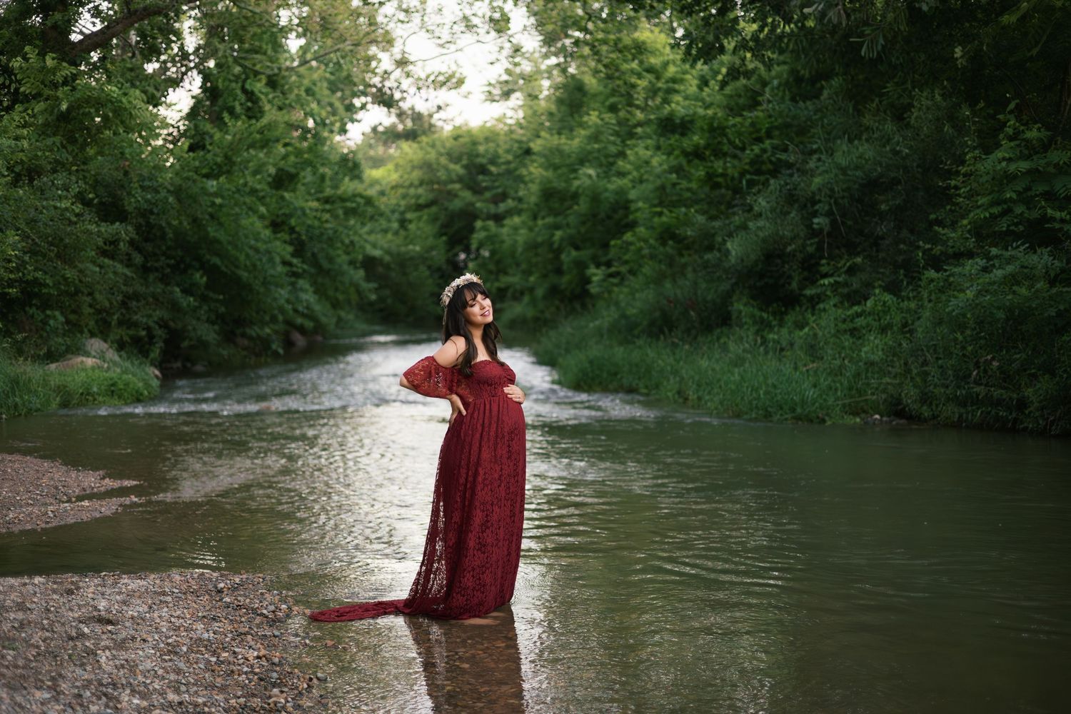 Woman in red lace gown standing in a shallow forest creek surrounded by lush greenery.