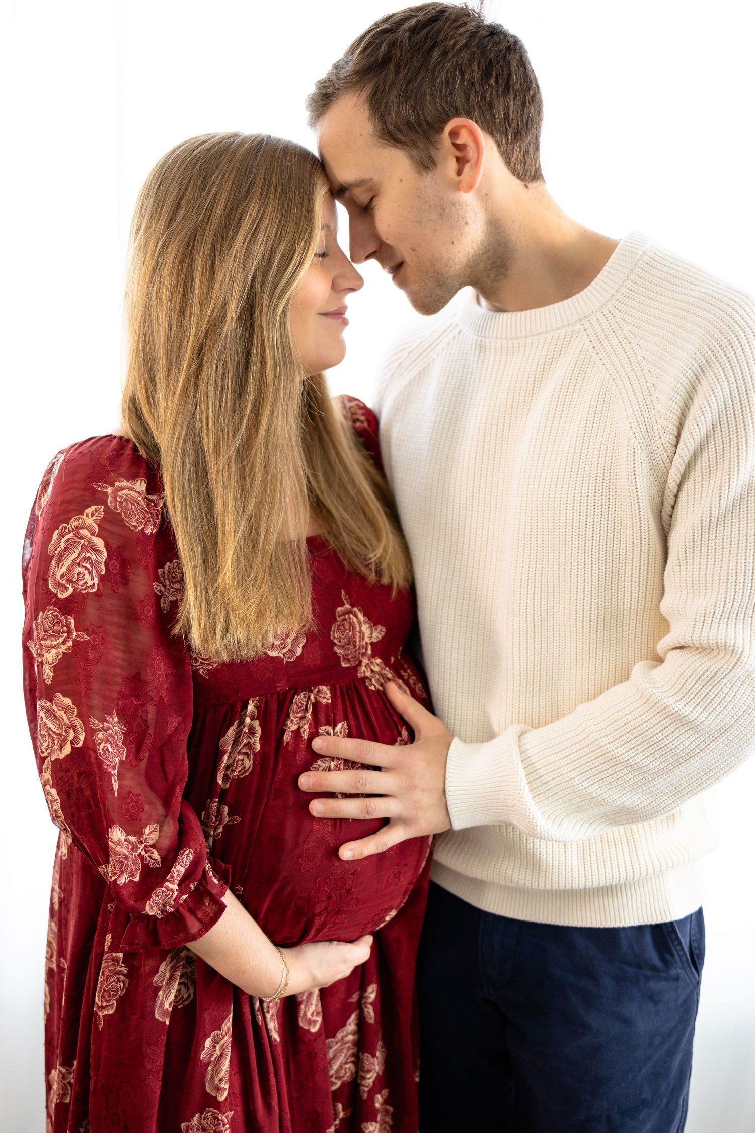 Romantic maternity portrait with woman in burgundy floral dress and man in white sweater during studio session in Hendricks County