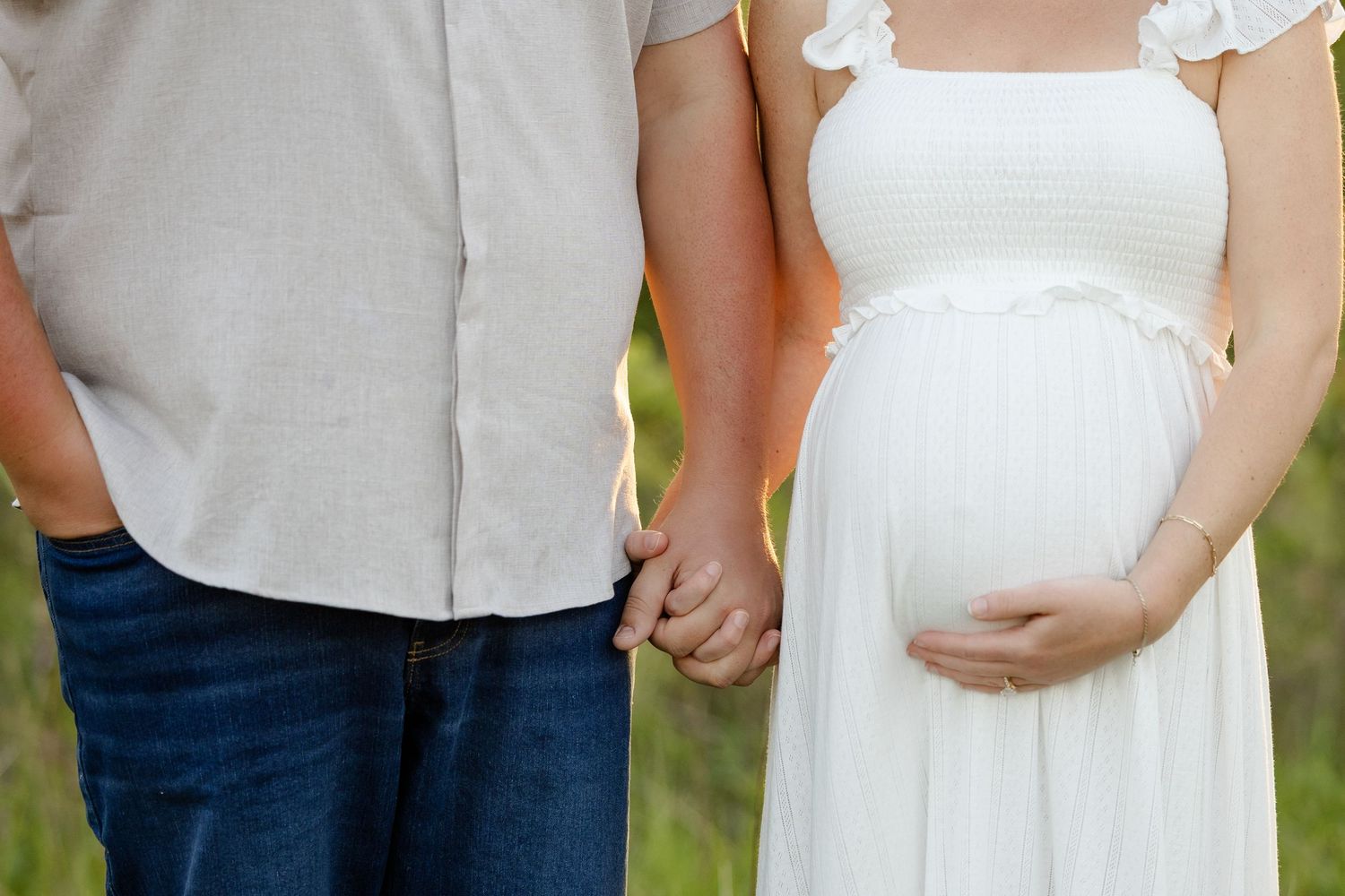 Pregnant woman in white dress holding hands with partner, cradling baby bump during Avon maternity photo session by Katy Rose Photography