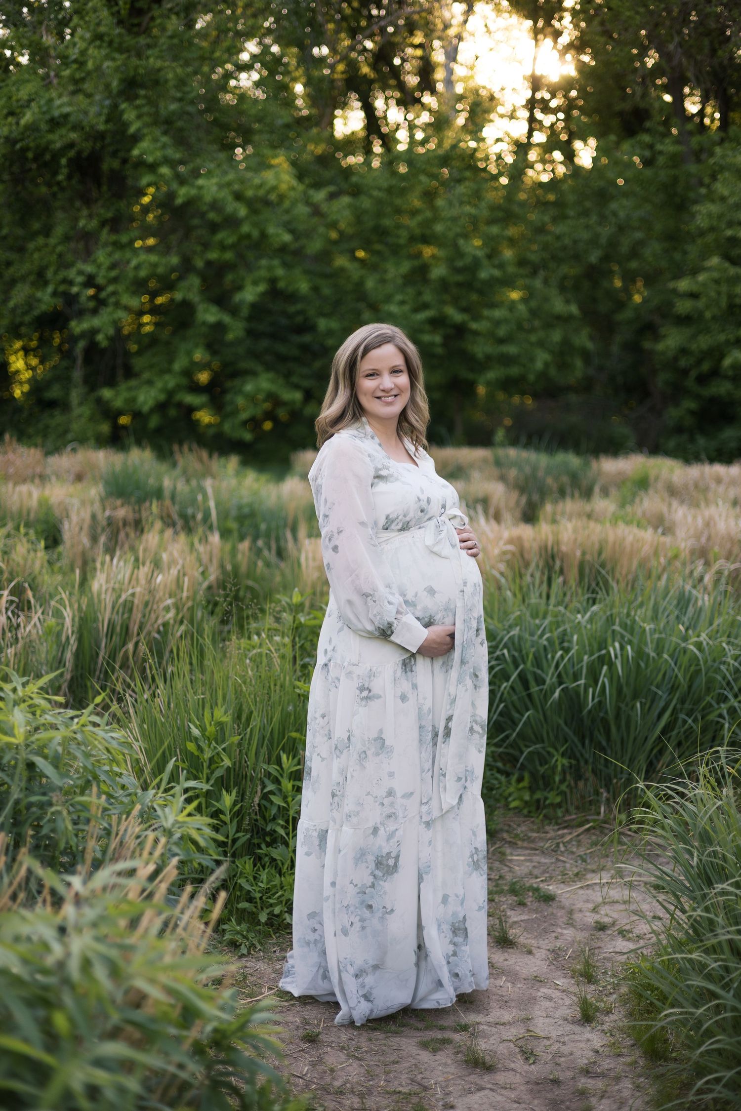 Smiling pregnant woman in floral maxi dress cradling baby bump outdoors in lush green field at golden hour.