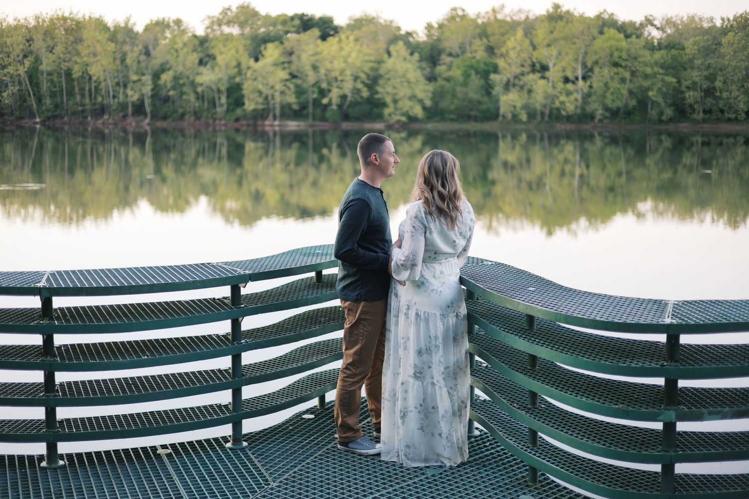 Couple standing together on dock surrounded by lush trees reflected in water for Indianapolis maternity session by Katy Rose Photography.