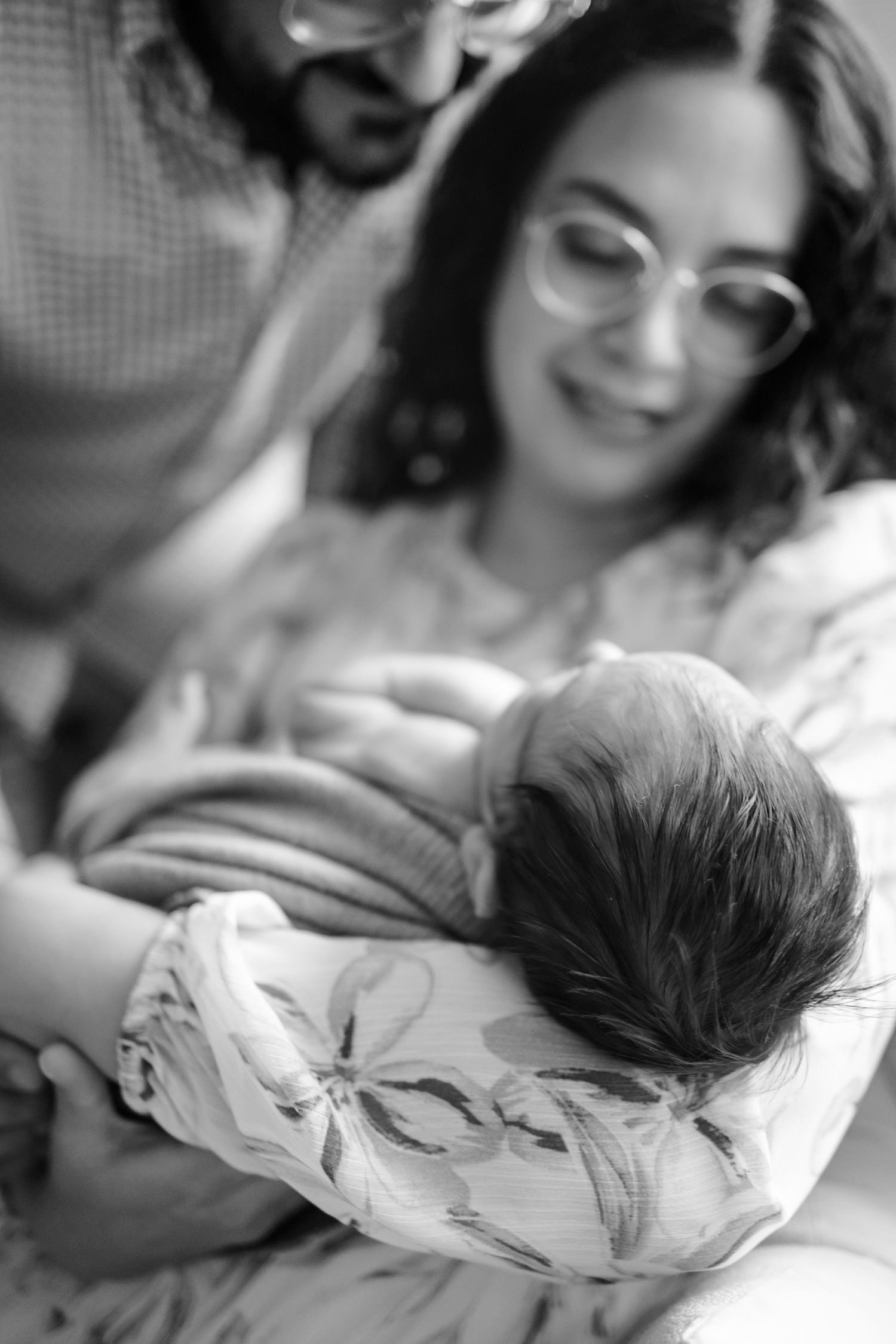 Black and white photo of parents lovingly gazing at their newborn baby wrapped in a floral swaddle.
