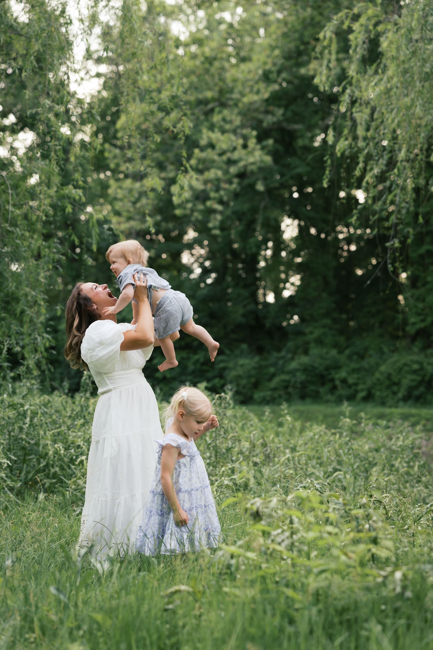 A mother in a white dress lifts her baby in a green park while her young daughter plays nearby.