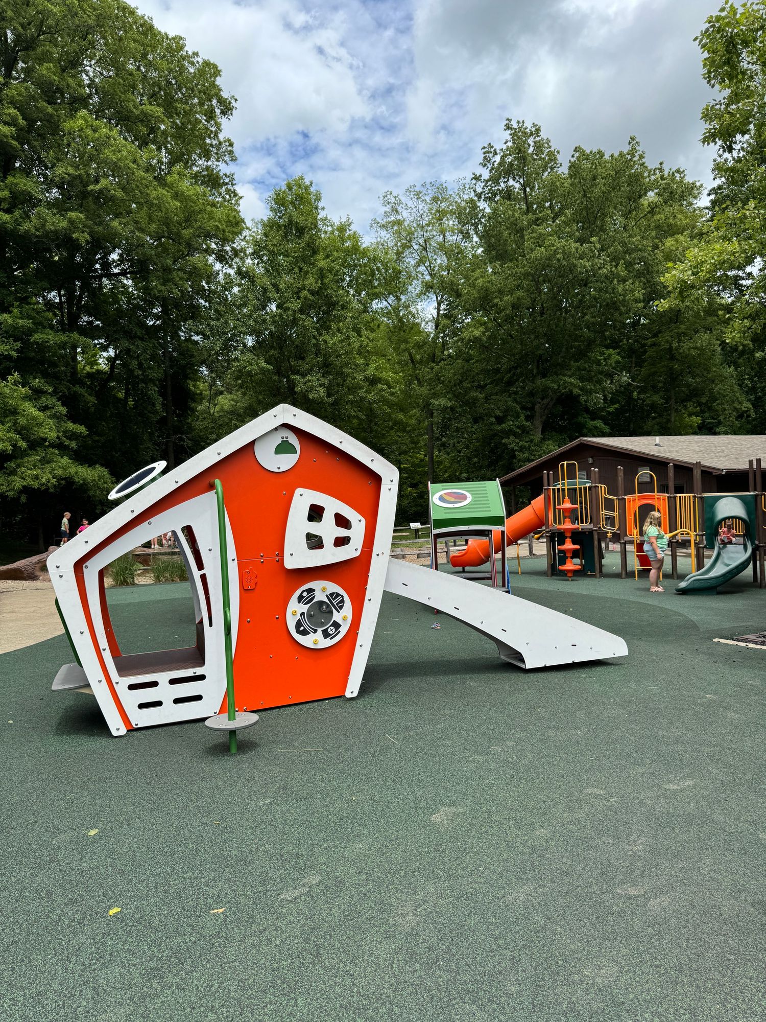 Colorful tilted playhouse with orange and white design on rubber playground surface surrounded by trees at Washington Township Park in Avon.