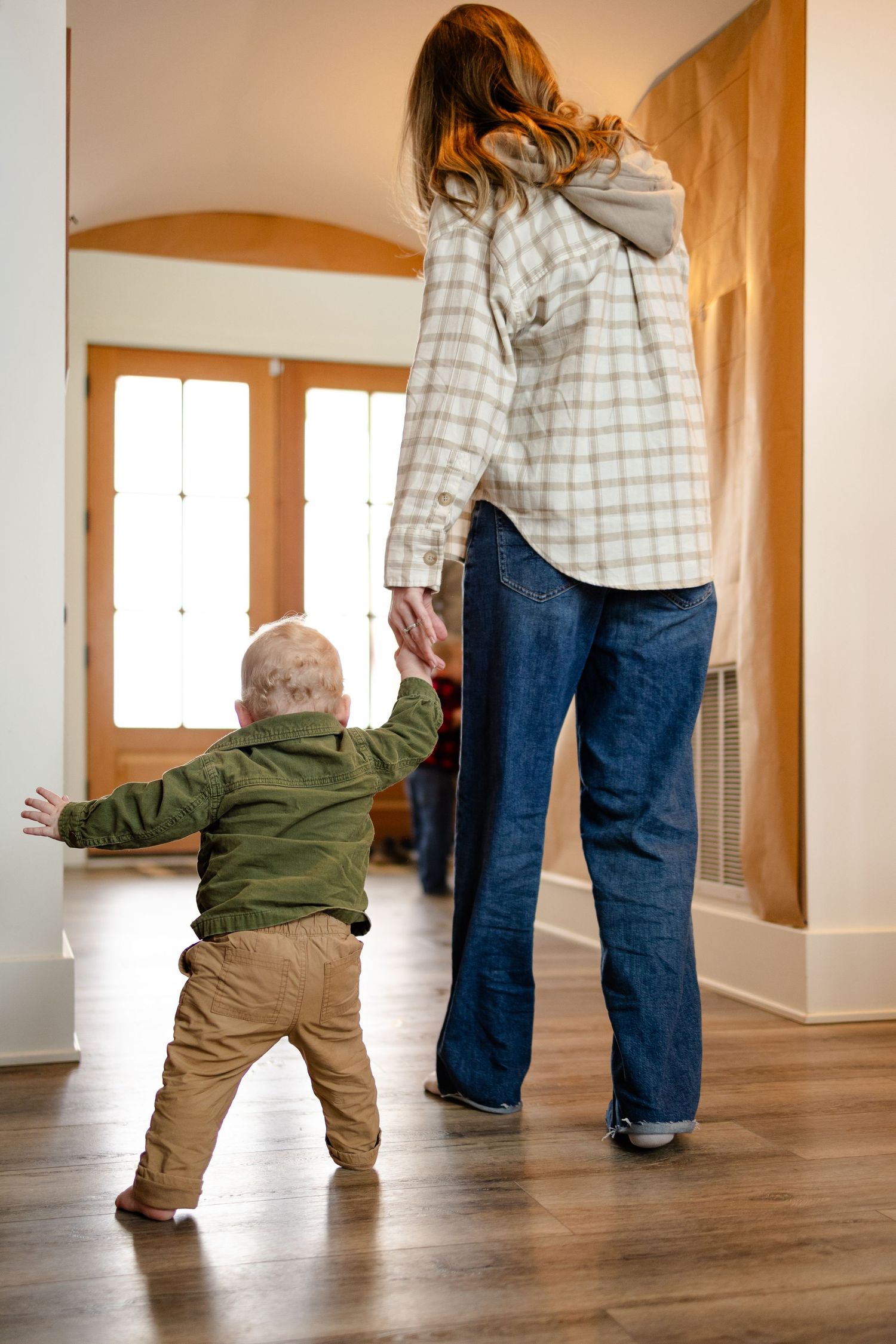 A toddler in khaki pants holds hands with his mom while walking on hardwood floors during lifestyle in-home session in Whitestown, Indiana