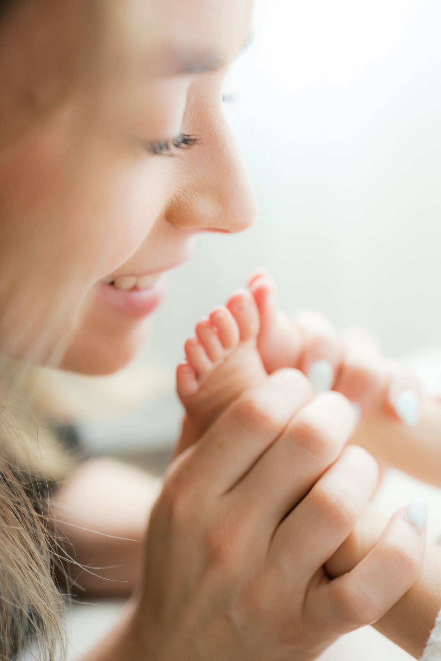 A smiling mother tenderly holds her newborn baby's tiny feet in her hands in a soft, bright setting.