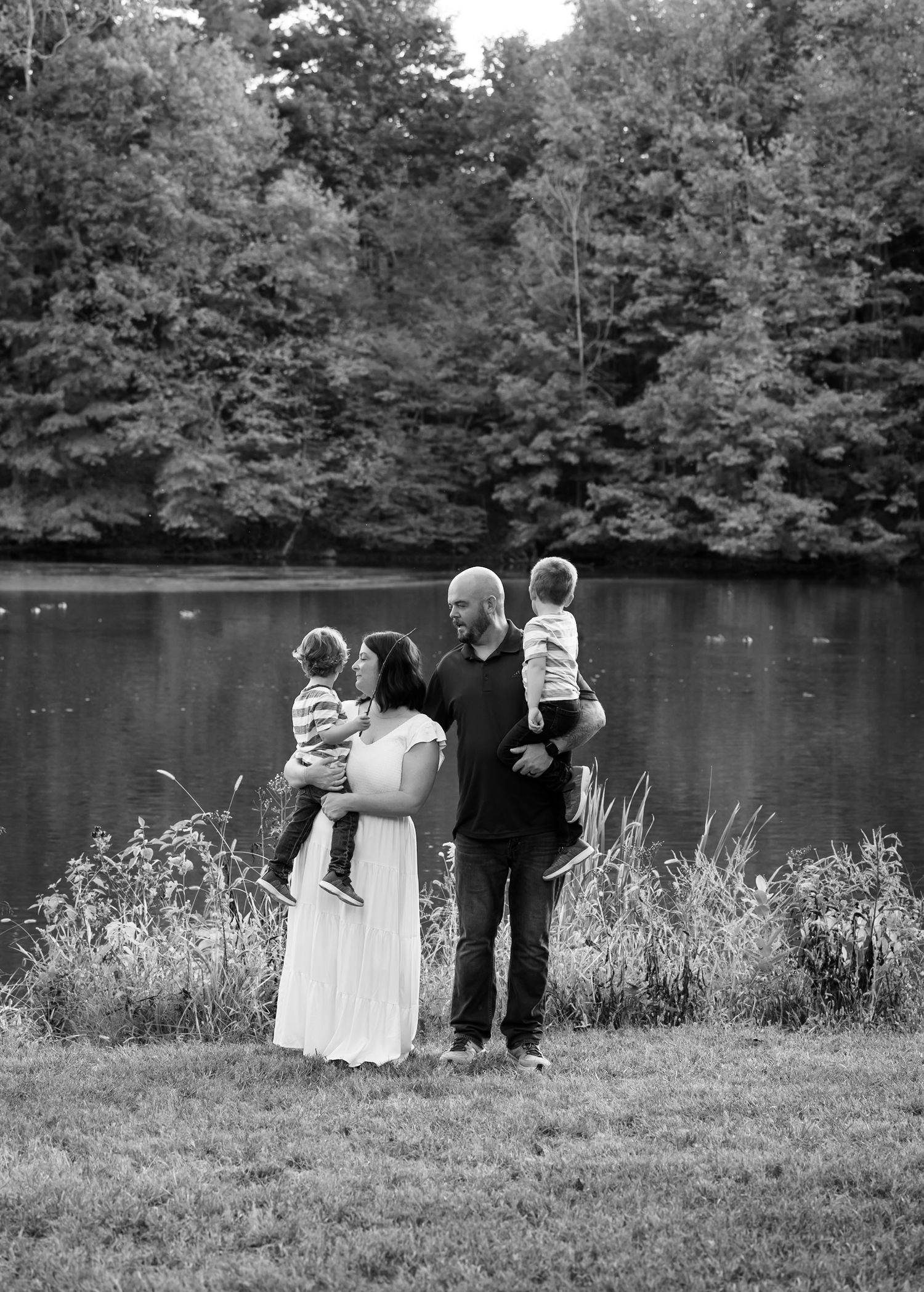 Three small figures stand by a tranquil lake surrounded by autumn trees in a black and white landscape.