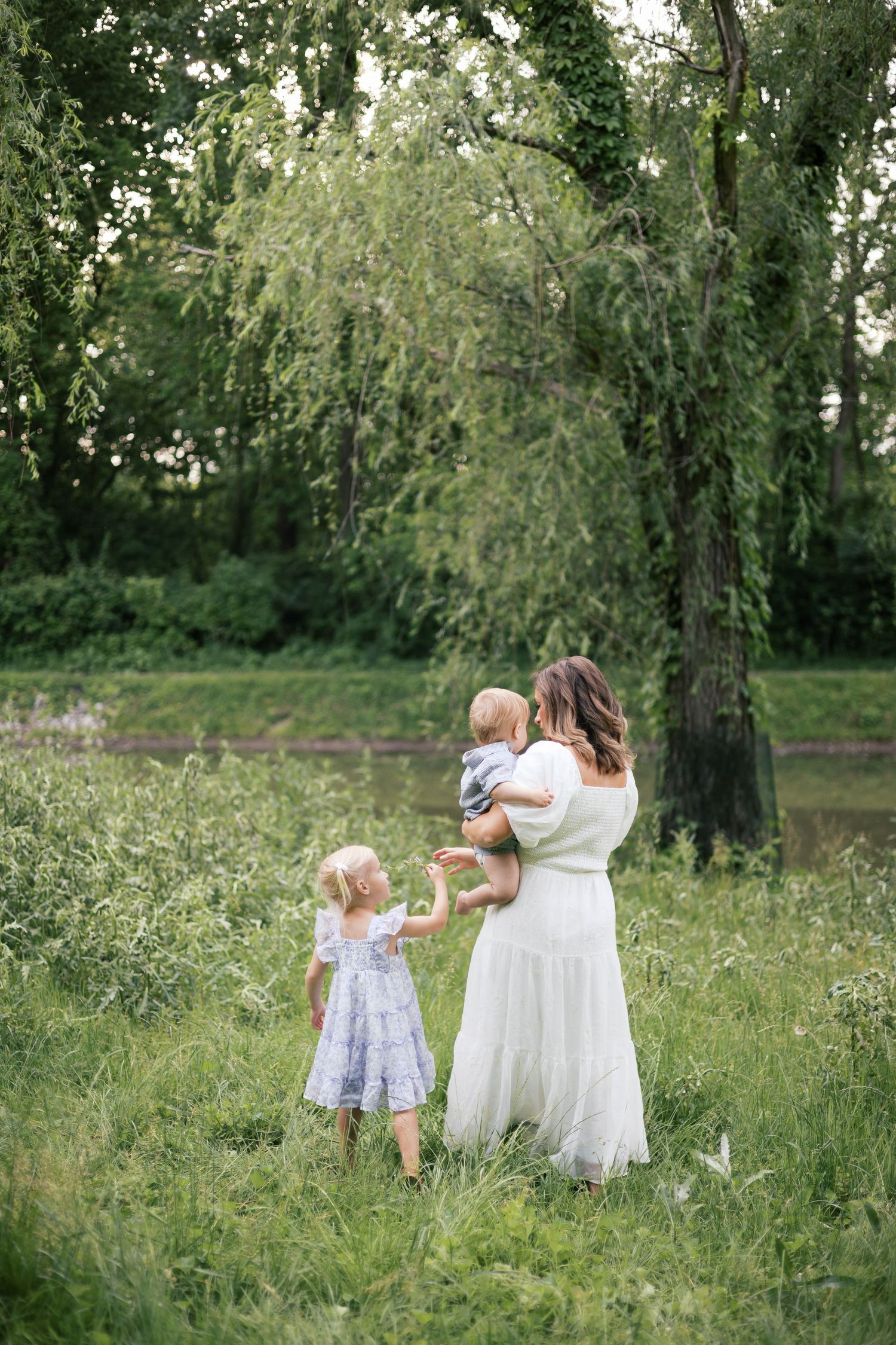 A mother in a white dress holds her baby while her young daughter in a blue dress stands beside her in a lush green park for family photos.