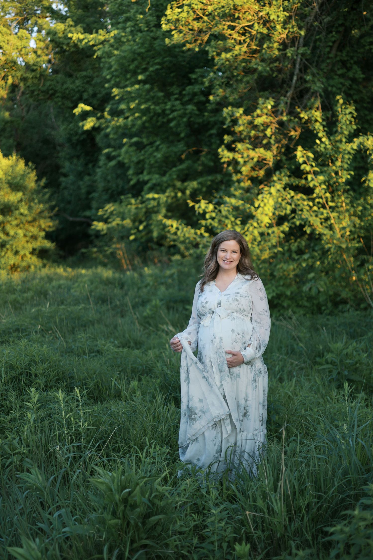 Smiling pregnant woman in a floral maxi dress standing in a lush green meadow at golden hour for Indianapolis Maternity Portraits.