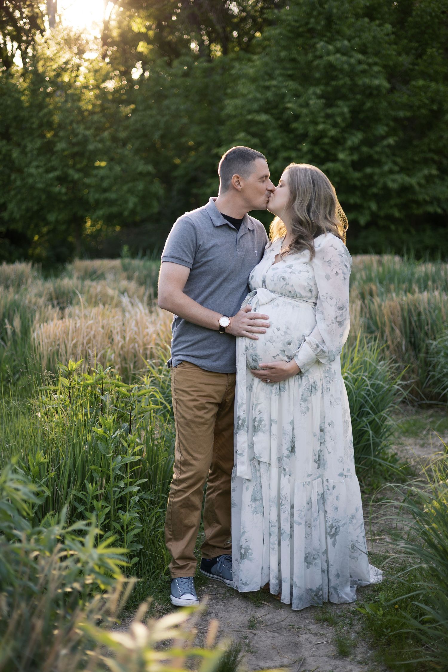 Couple sharing a kiss outdoors at sunset, pregnant woman in floral dress with partner in gray shirt, surrounded by lush greenery.