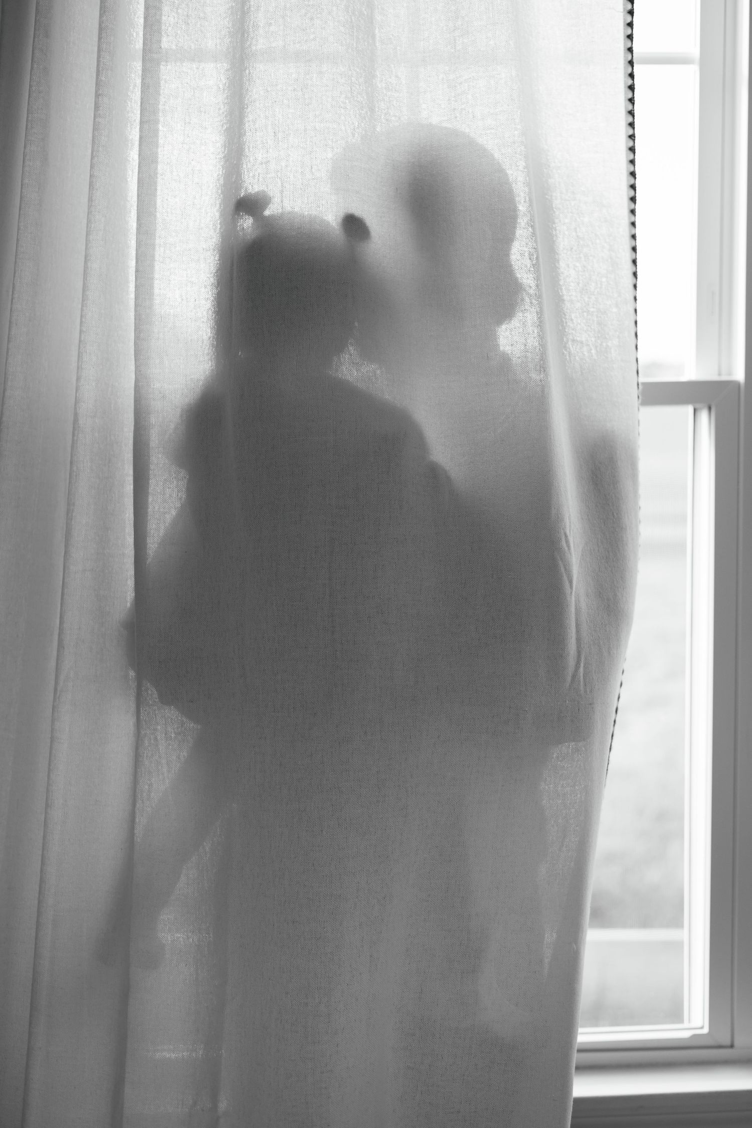 Silhouette of mother and child behind sheer curtain near window in black and white photography.