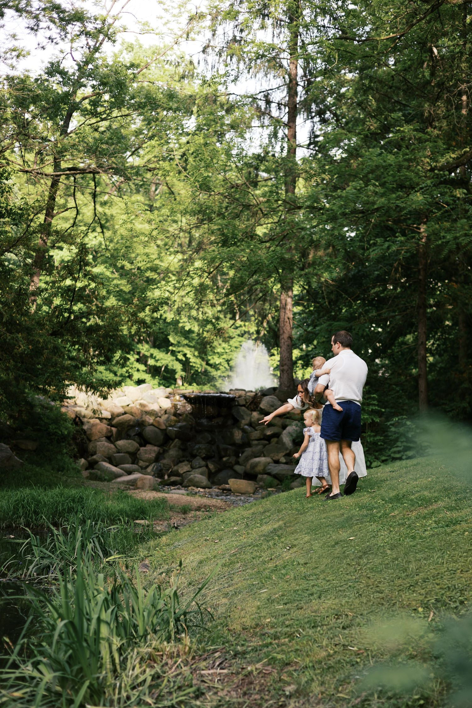 Family of four admiring a stone waterfall fountain surrounded by lush green trees in a park.