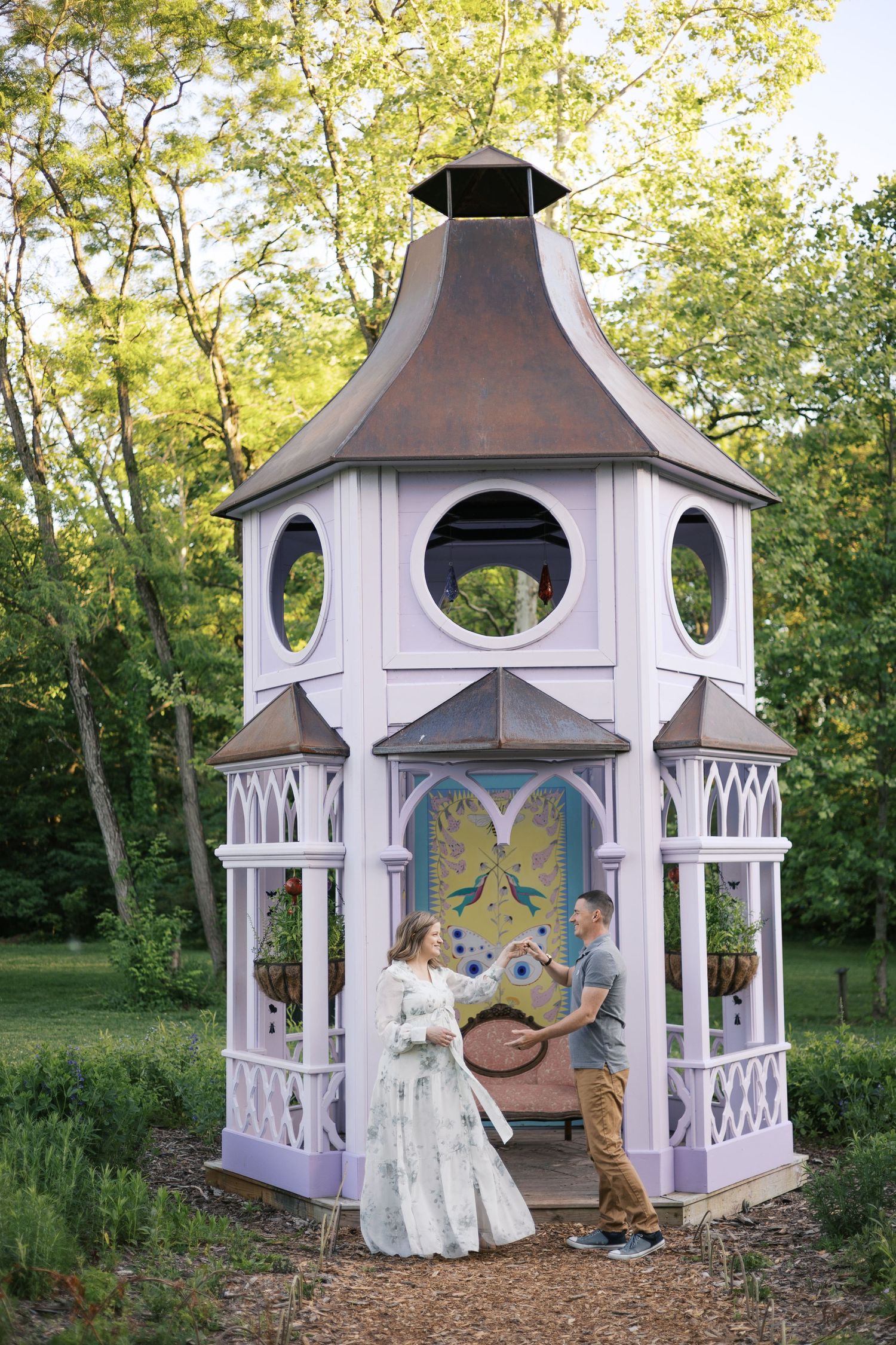 Pregnant couple sharing a tender moment beside a charming lavender gazebo surrounded by lush green trees.
