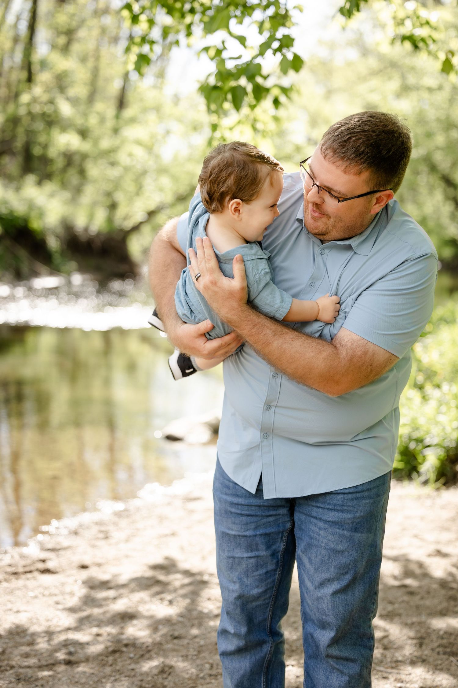 Father holding baby boy near a creek surrounded by lush green trees on a sunny day at Arbuckle Park in Brownsburg, Indiana.