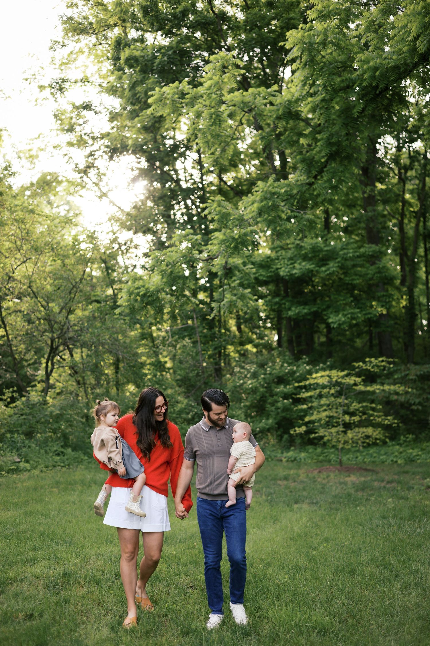 A young family walks hand-in-hand on green grass, each parent holding a toddler, surrounded by lush trees.