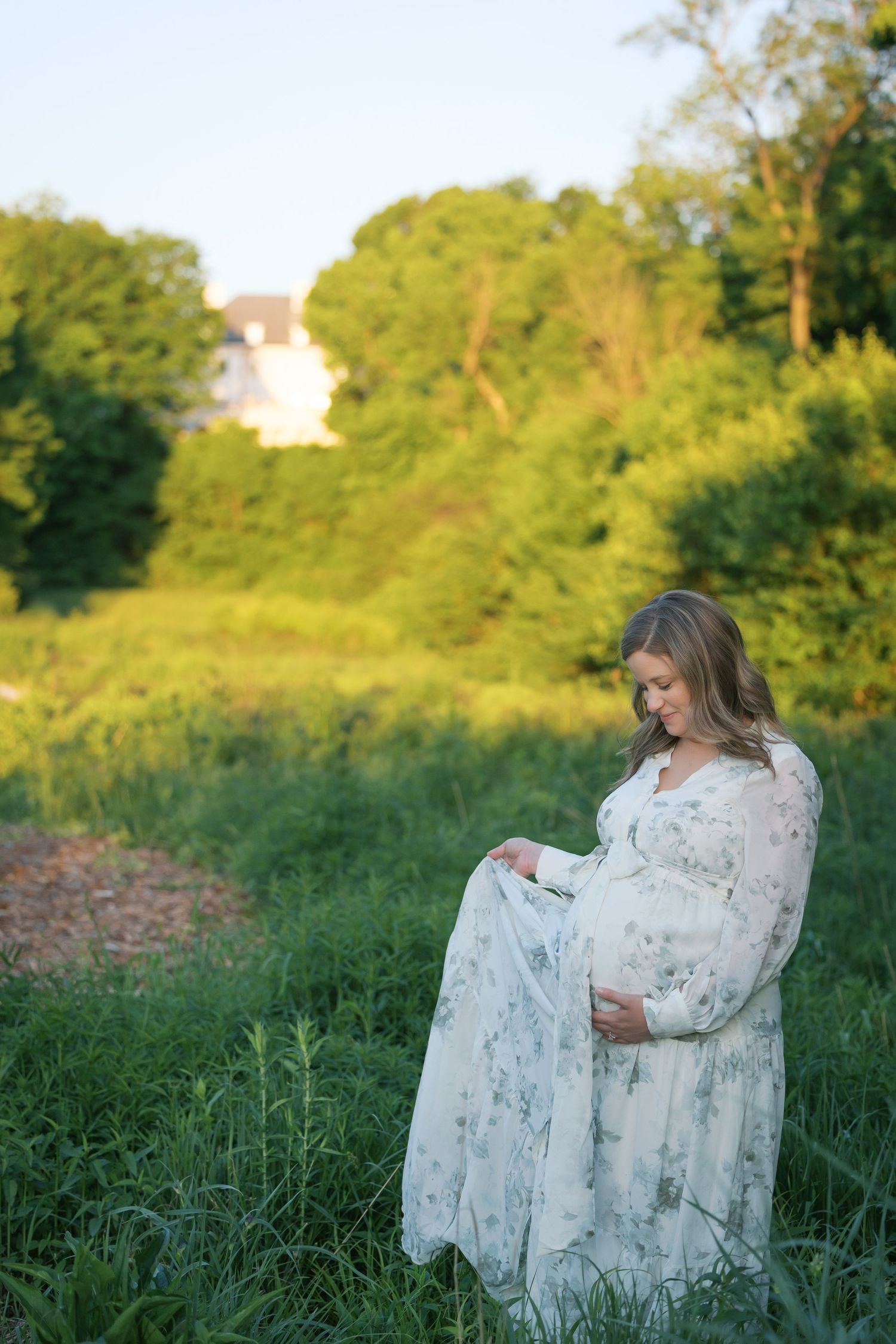 Pregnant woman in floral maxi dress standing in green field during golden hour maternity photoshoot.