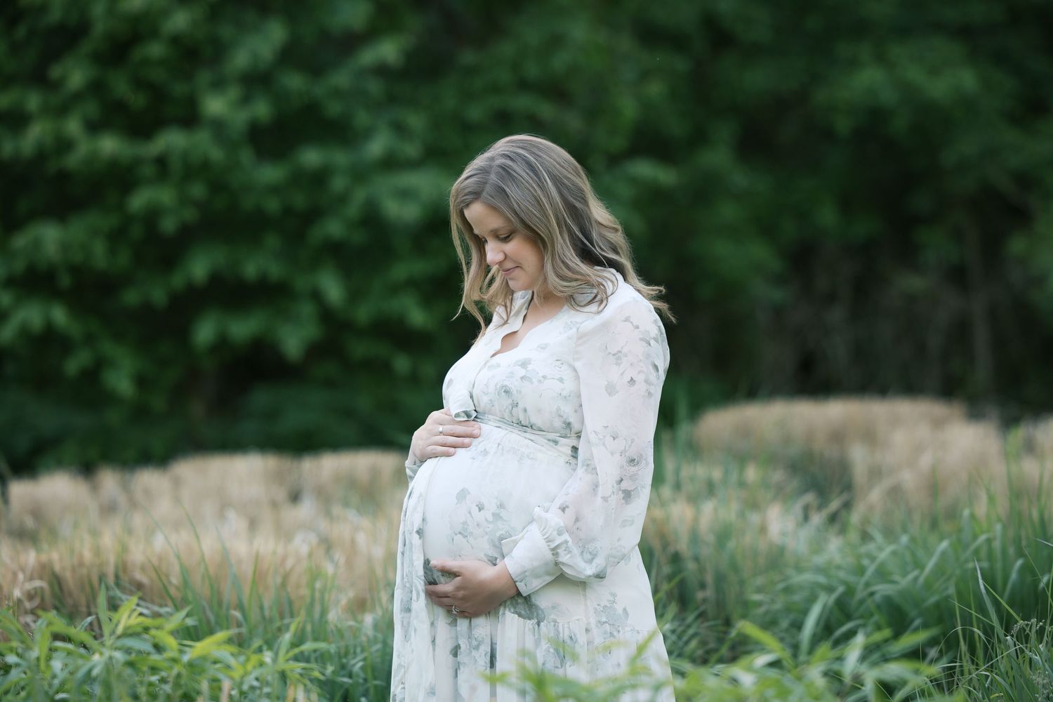 Pregnant woman in floral dress cradling baby bump in lush green field during outdoor maternity photo session by Katy Rose Photography.