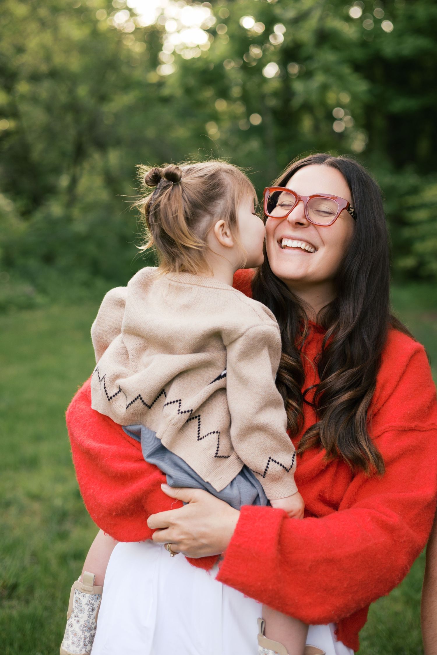 A laughing woman in a red sweater holds a young girl who kisses her cheek outdoors in a green park.