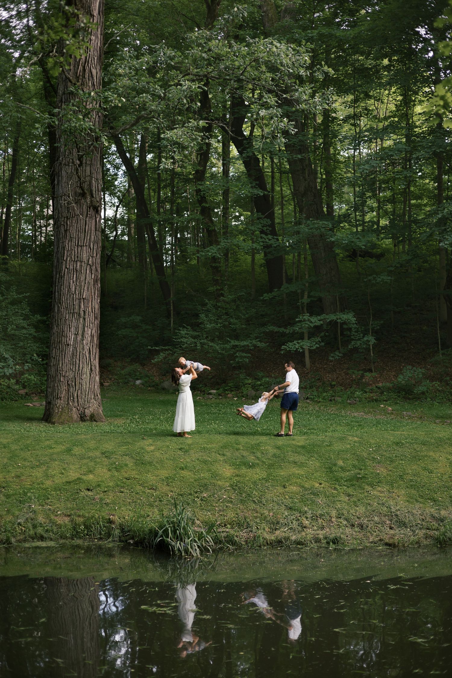 Family playing by a serene pond in a lush green forest, with reflections visible in the calm water below.