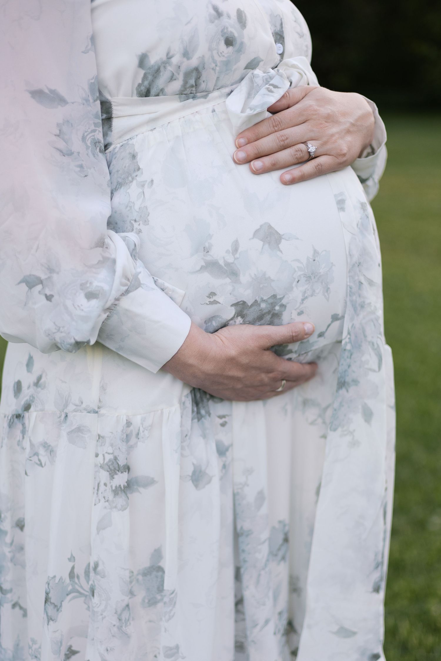 Close-up of pregnant woman cradling baby bump in elegant white floral maternity dress during maternity session by Katy Rose Photography.