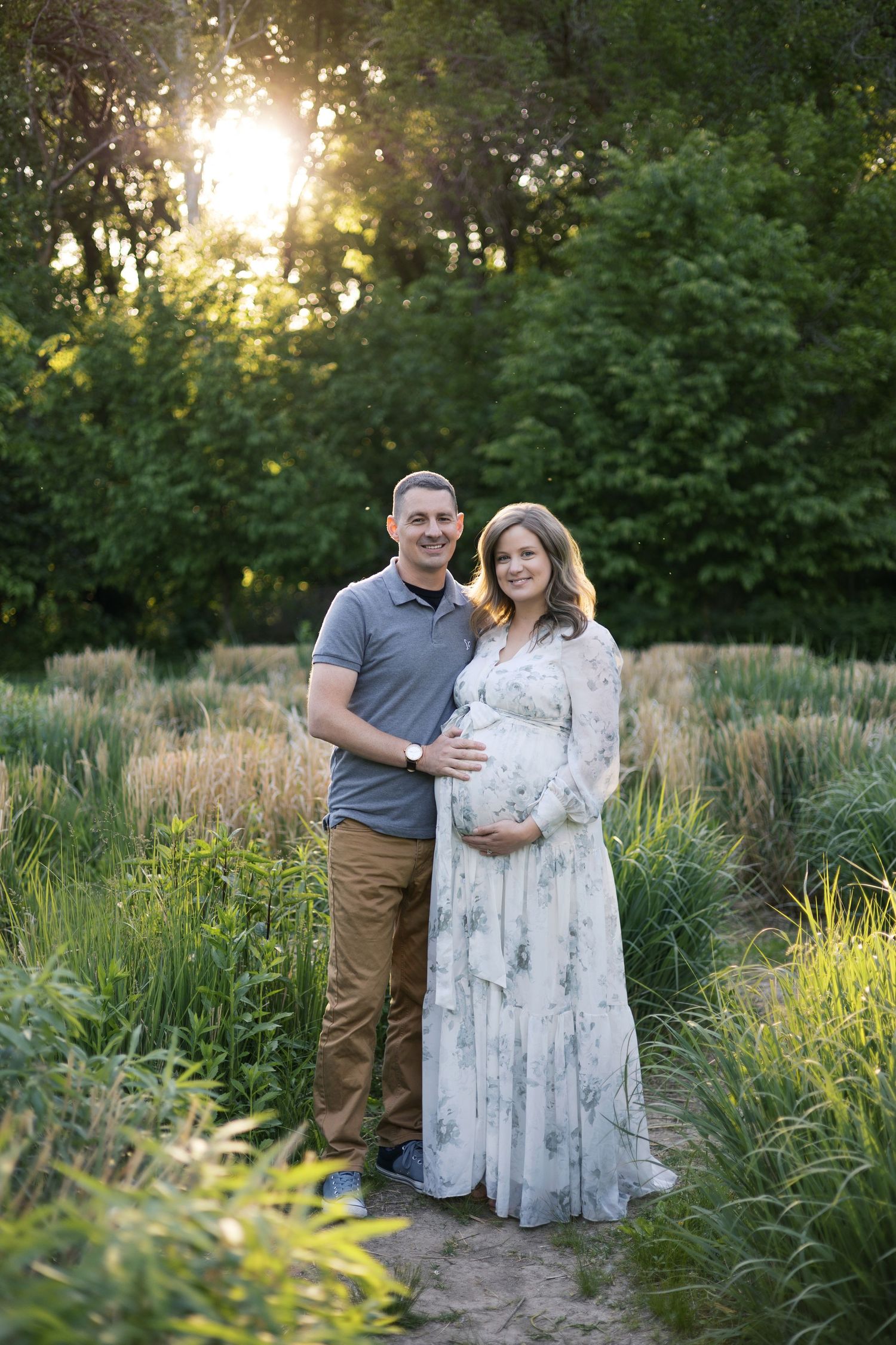 Couple poses in lush garden at golden hour, pregnant woman in floral maxi dress cradling baby bump during maternity photoshoot at Newfields
