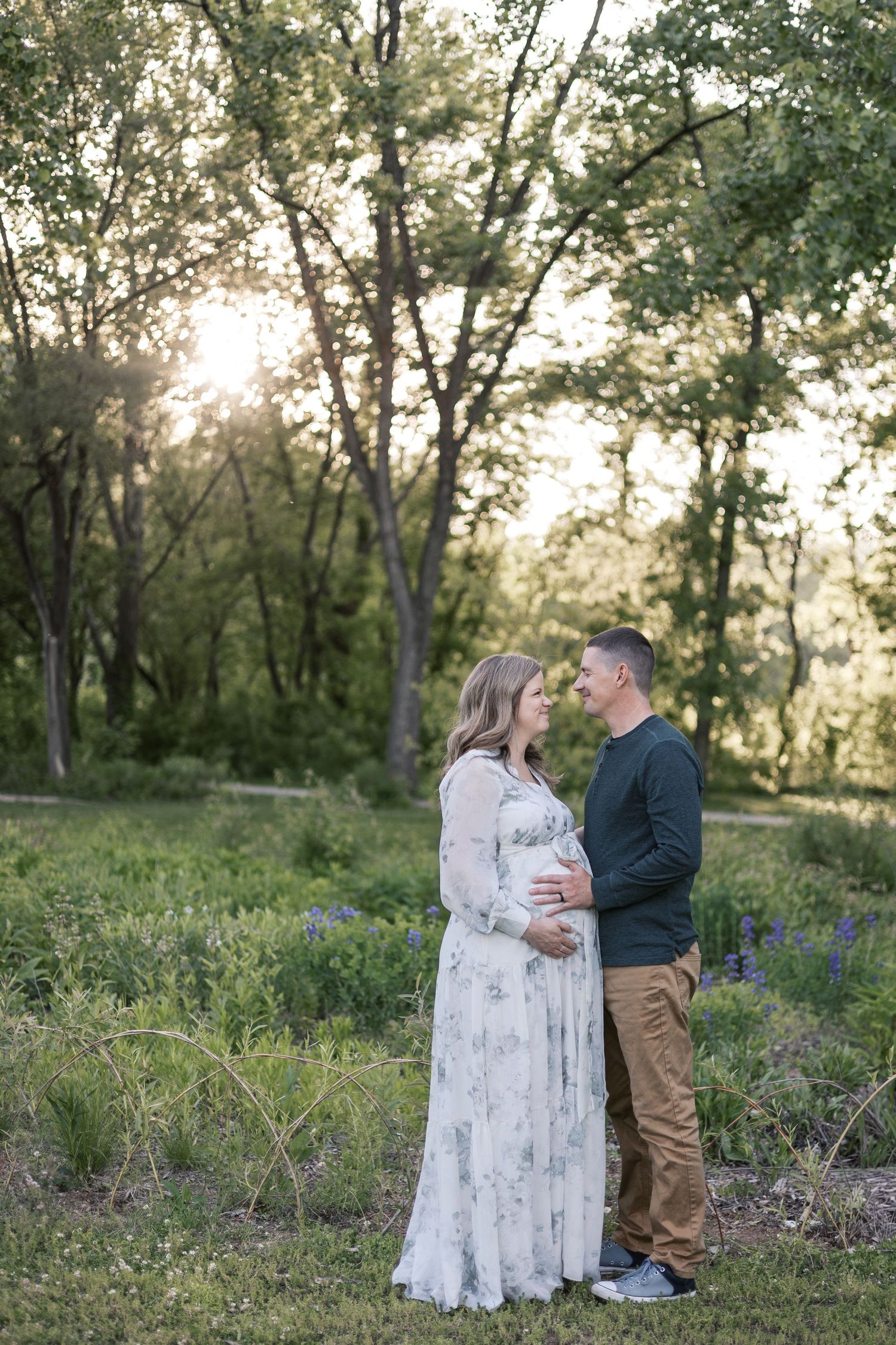 Pregnant couple embraces in wildflower meadow at sunset, woman in floral maternity gown, romantic outdoor portrait.