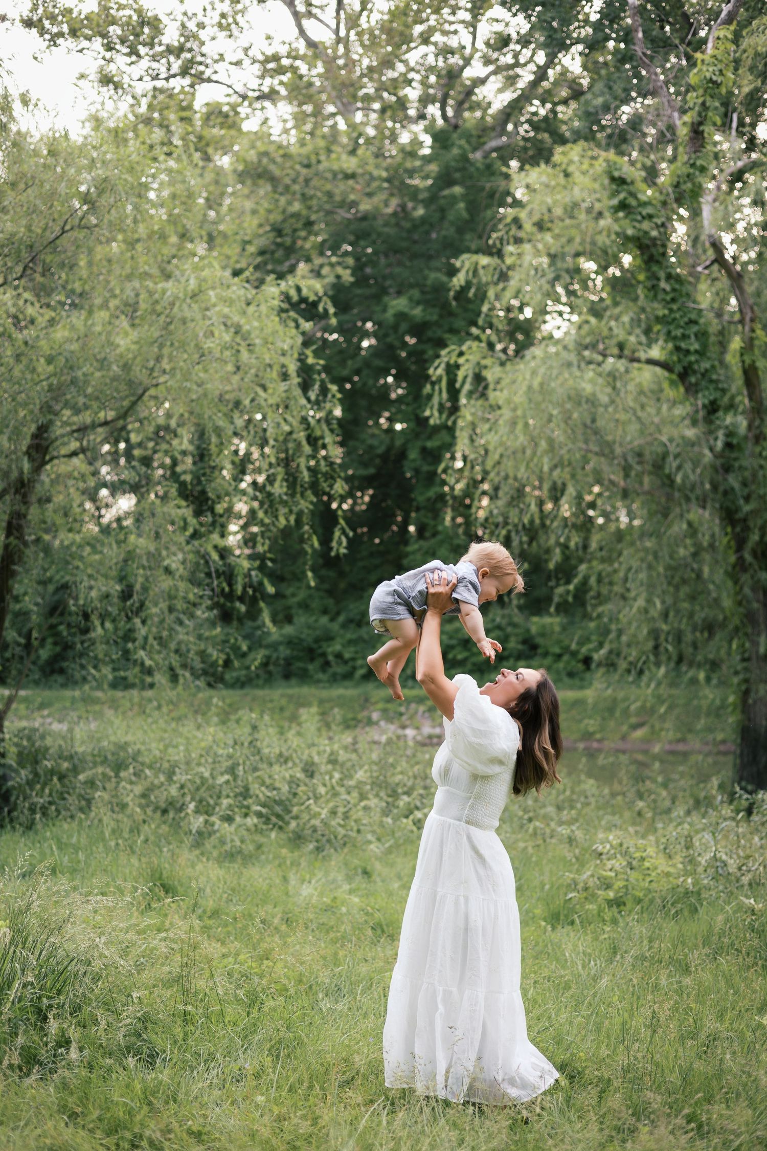 A smiling mother in a white dress lifts her baby joyfully in a green outdoor park setting.