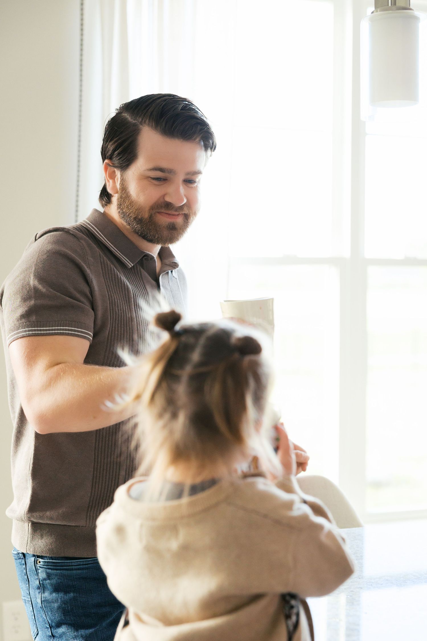 A smiling bearded man brushing a young child's hair near a bright window indoors.