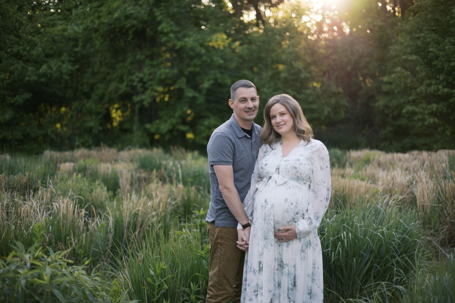 Couple poses in sunlit field, pregnant woman in floral dress holding belly with partner during maternity photo session.