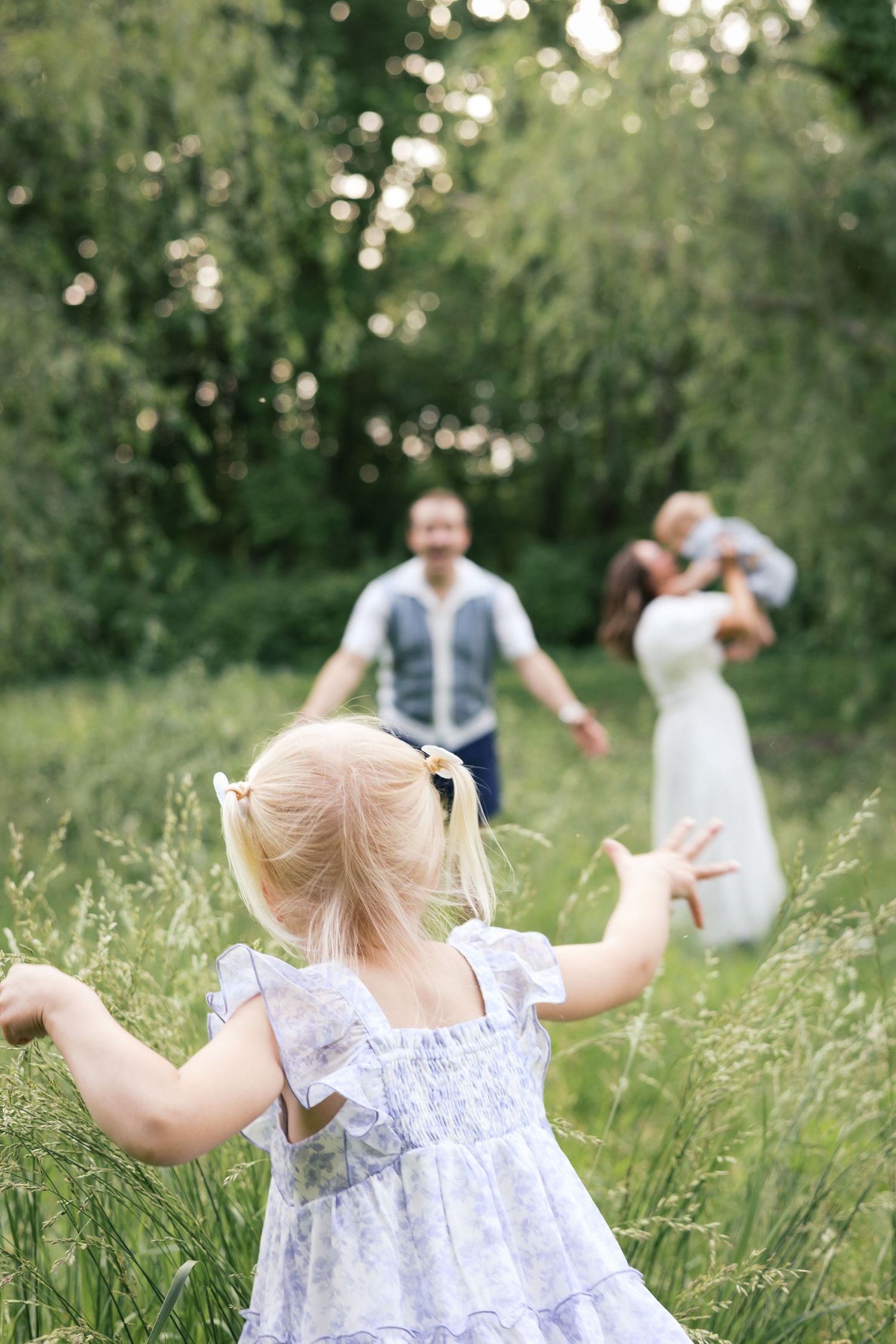 A young blonde girl in a floral dress runs toward her family in a lush green meadow on a sunny day.