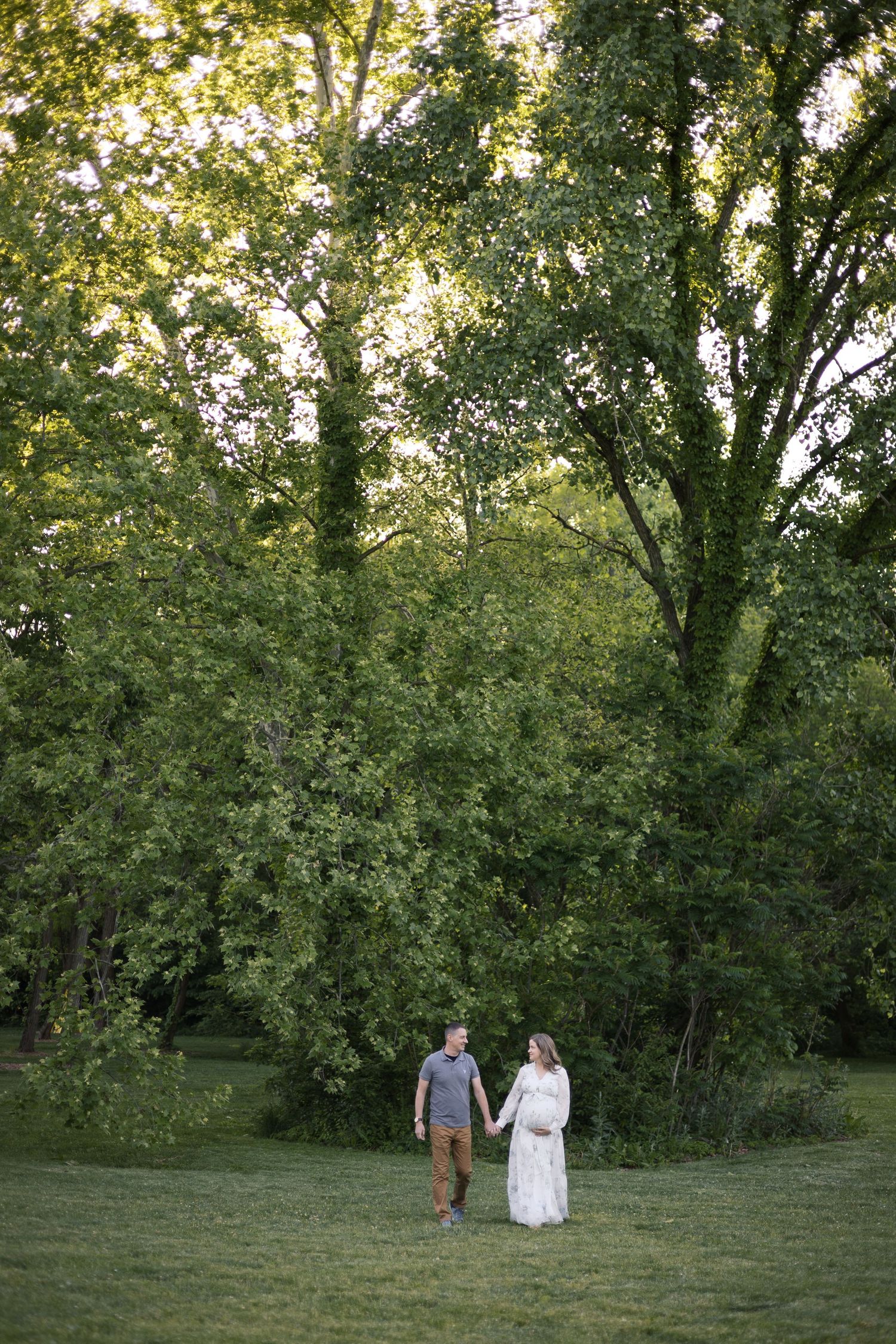 Expecting couple holds hands walking in green park, woman in white maternity dress, golden hour outdoor photography.