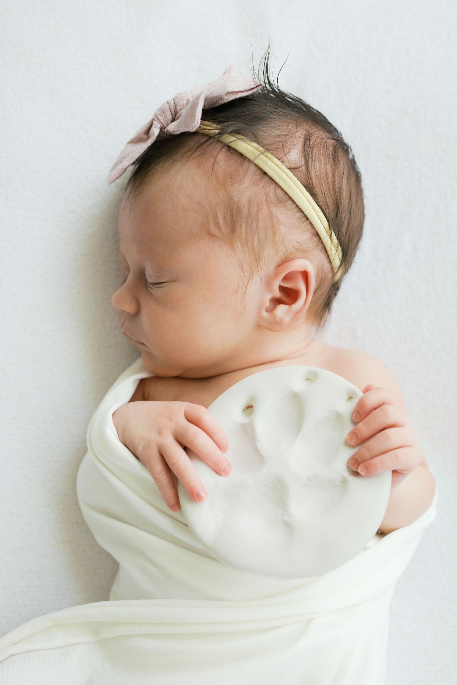 Sleeping newborn baby girl swaddled in white wrap wearing a pink bow headband, lying on a white background.