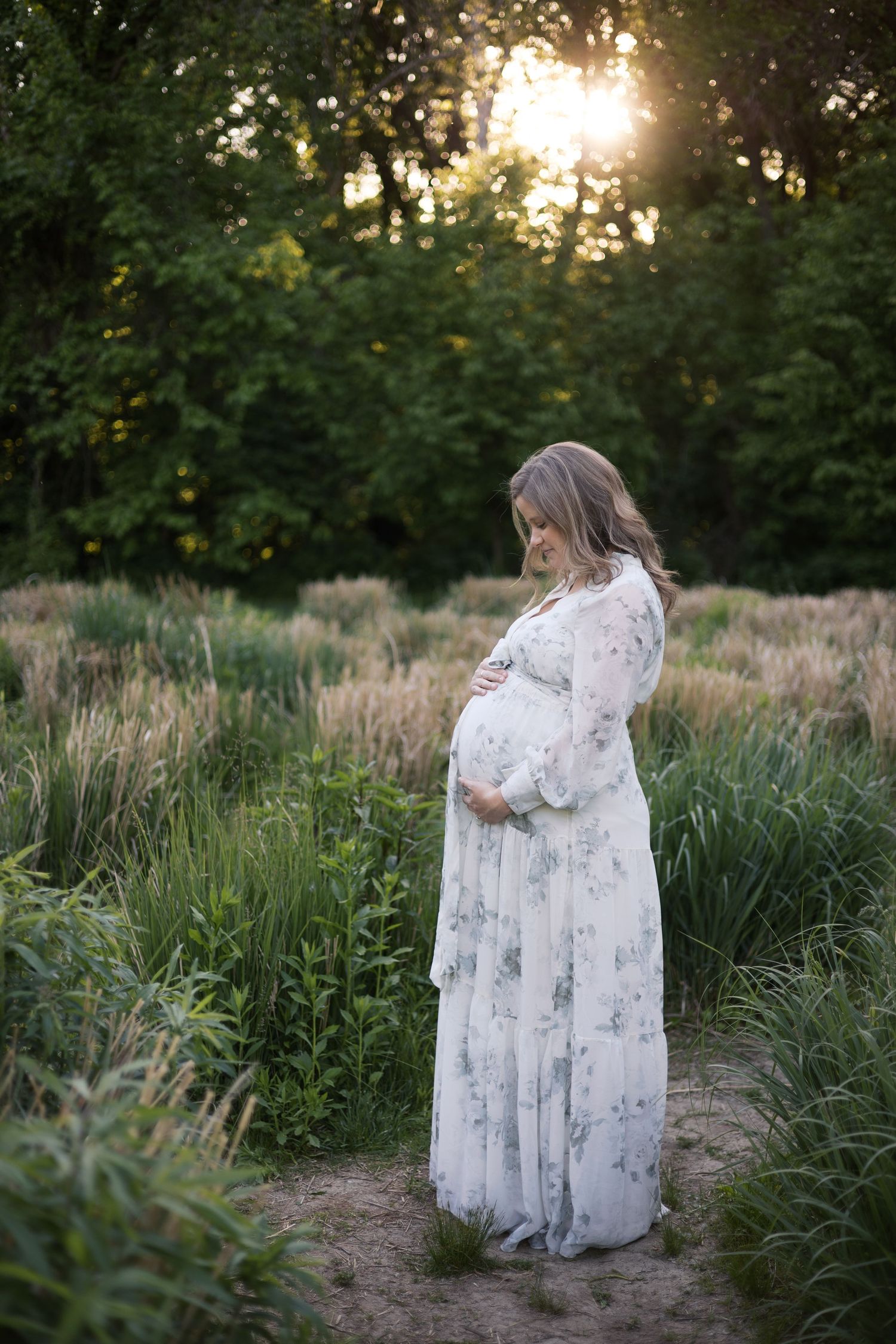 Pregnant woman in floral maxi dress cradling baby bump in lush green field at golden hour for maternity photos by Katy Rose Photography.