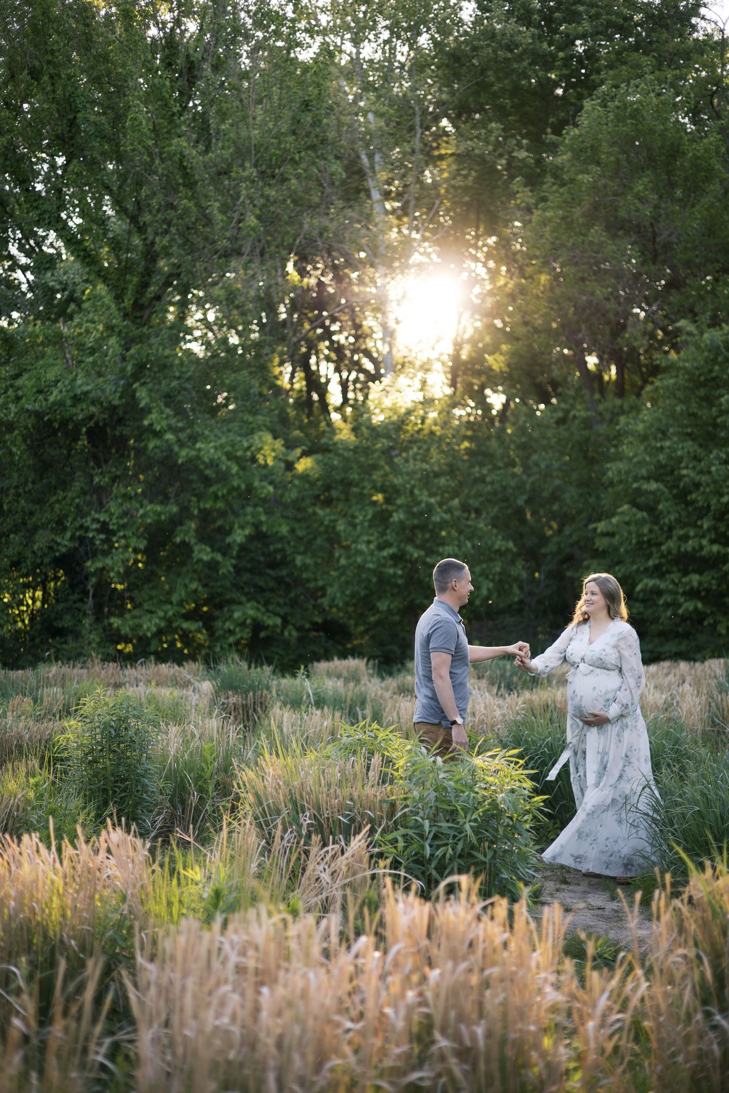 Pregnant woman in white gown holds hands with partner in golden hour sunlight among tall grasses and lush green trees.