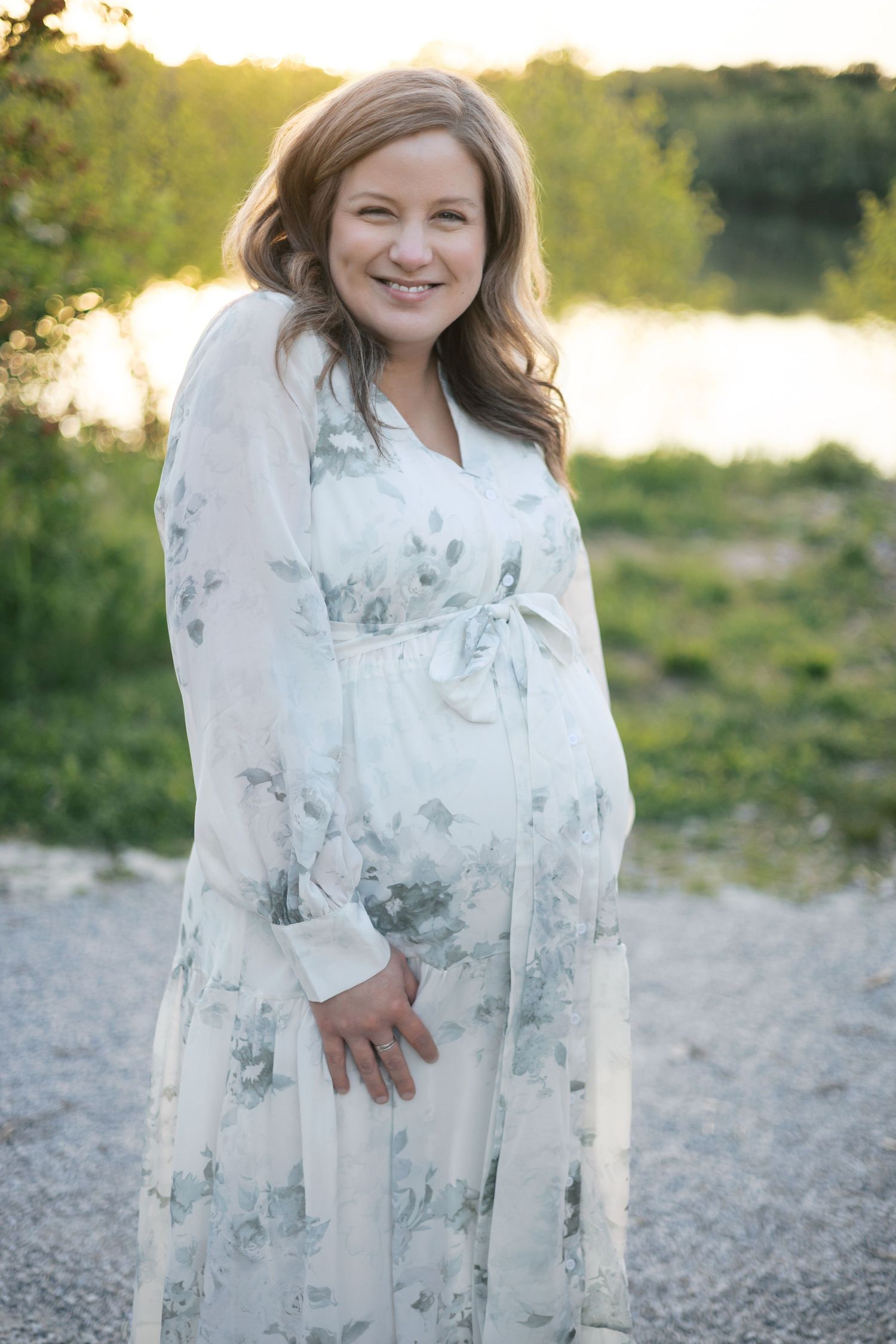 Smiling pregnant woman in white floral dress standing by lake in for Newfield's maternity session by Katy Rose Photography.