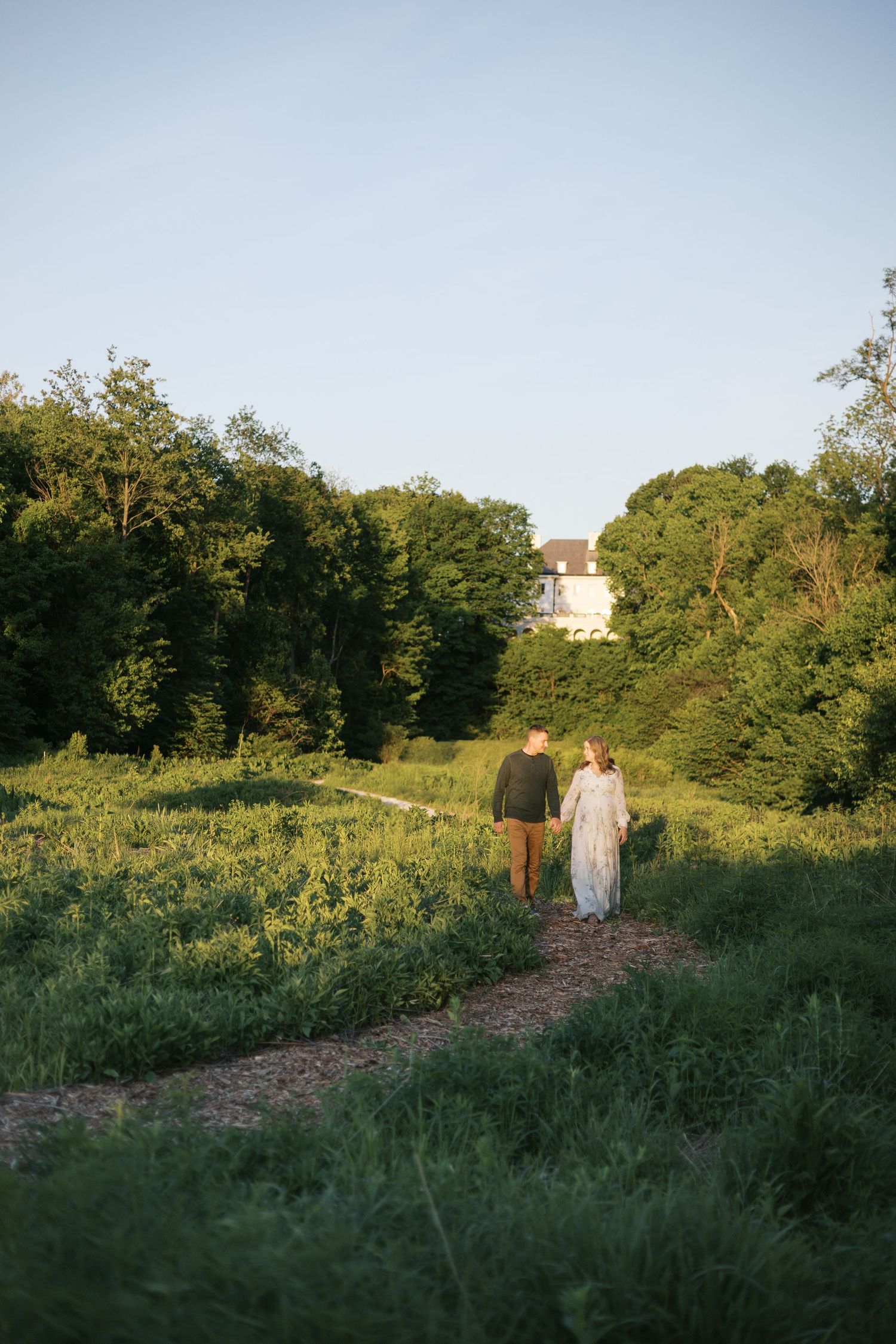 Couple holding hands walking along a garden path surrounded by lush green trees and fields at golden hour during maternity photos