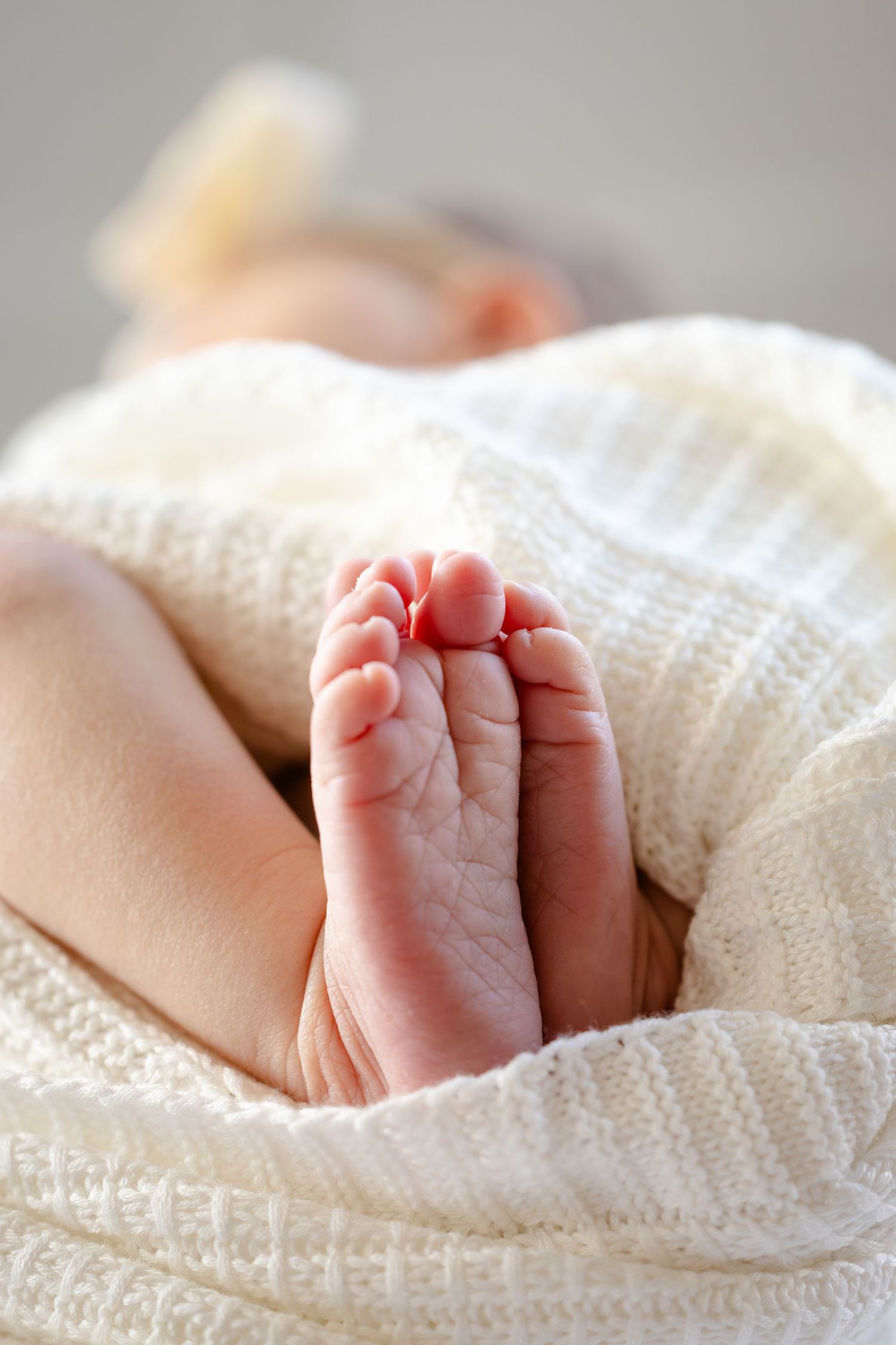 Tiny baby feet wrapped in soft white knitted blanket showing delicate toes in gentle natural lighting during in-home newborn session