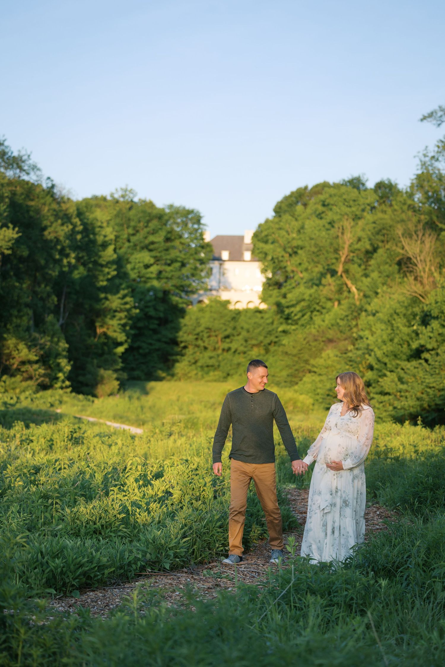 Couple holding hands in a sunlit green meadow, pregnant woman in floral dress, white house visible through trees.