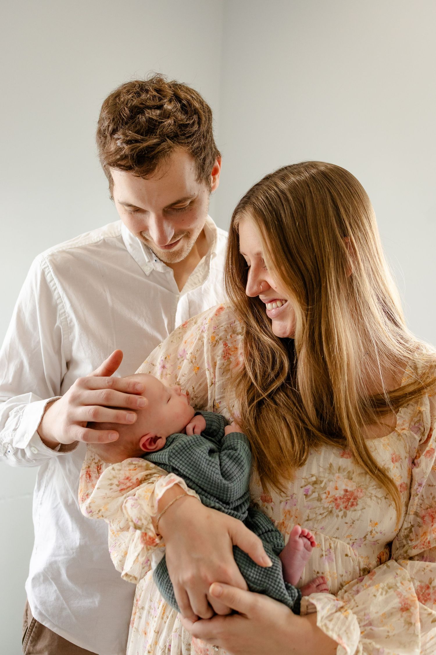 A couple in white and floral attire share intimate moments while holding their newborn baby in a bright room during newborn session
