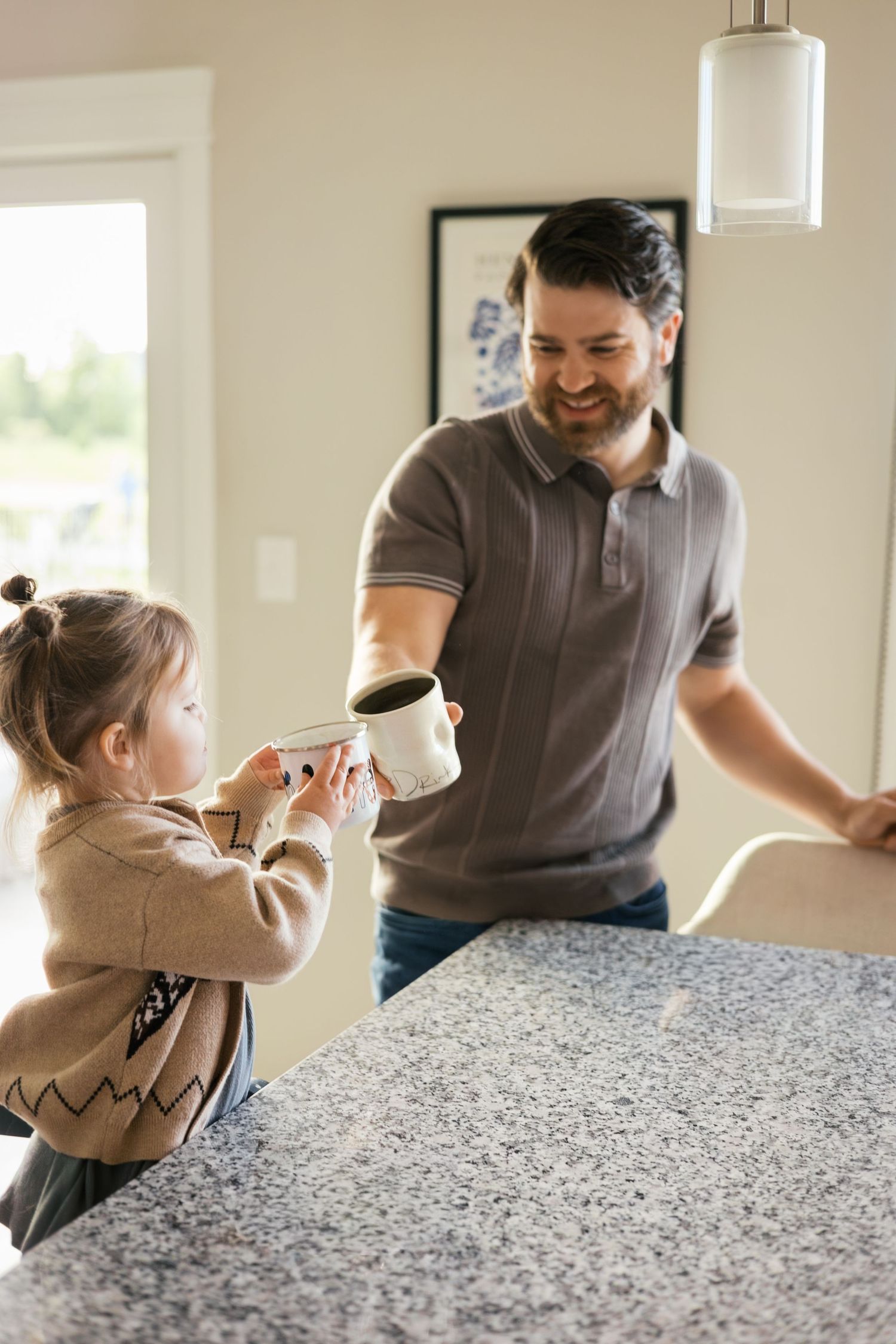 A smiling father hands a coffee mug to his young daughter in a bright modern kitchen.