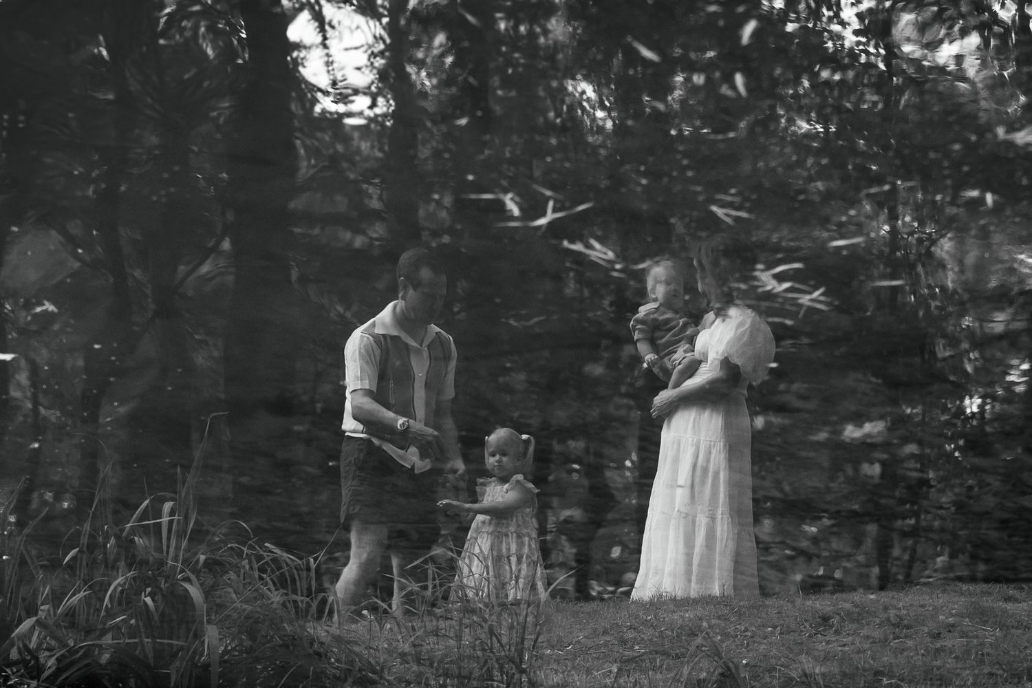 Black and white photo of a family with a toddler and pregnant mother standing among trees outdoors.
