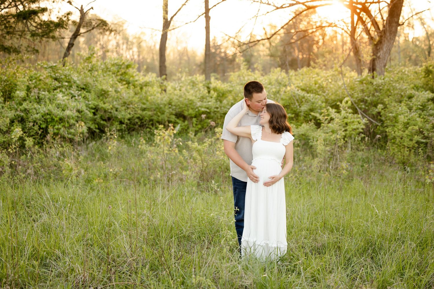Couple sharing a tender moment in a sunlit meadow, man kissing pregnant woman in white maternity dress at golden hour.